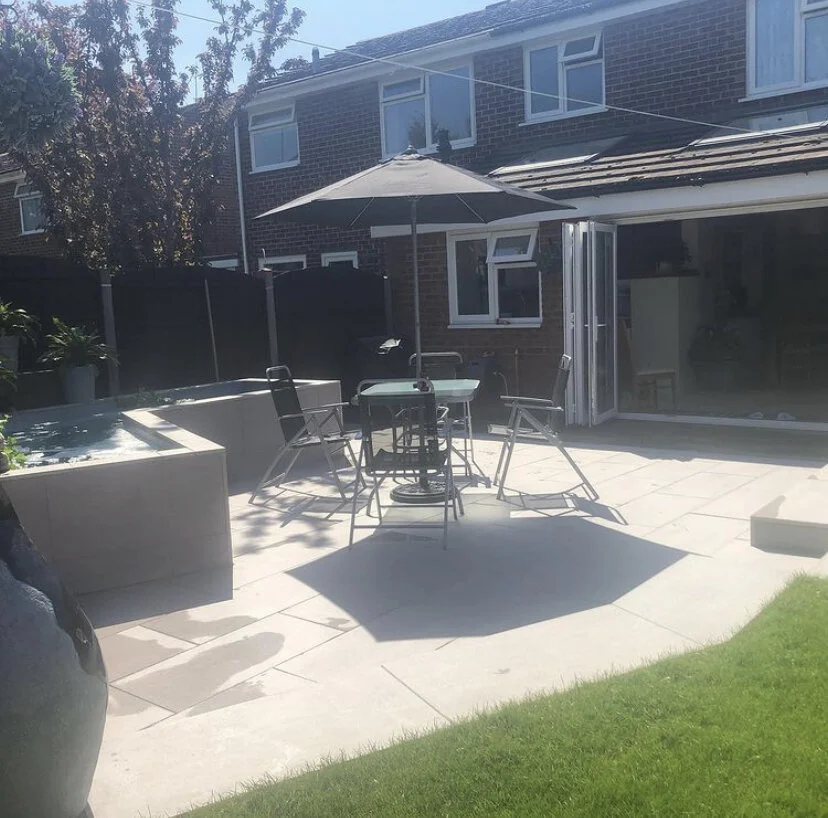 Backyard patio area with a hot tub, outdoor dining table with chairs, and a large umbrella, adjacent to a brick house with a sliding glass door and several windows, on a sunny day.