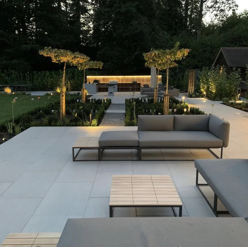 Modern outdoor patio with gray cushioned seating, small wooden tables, and a garden with trees and plants, illuminated by soft lighting at dusk.