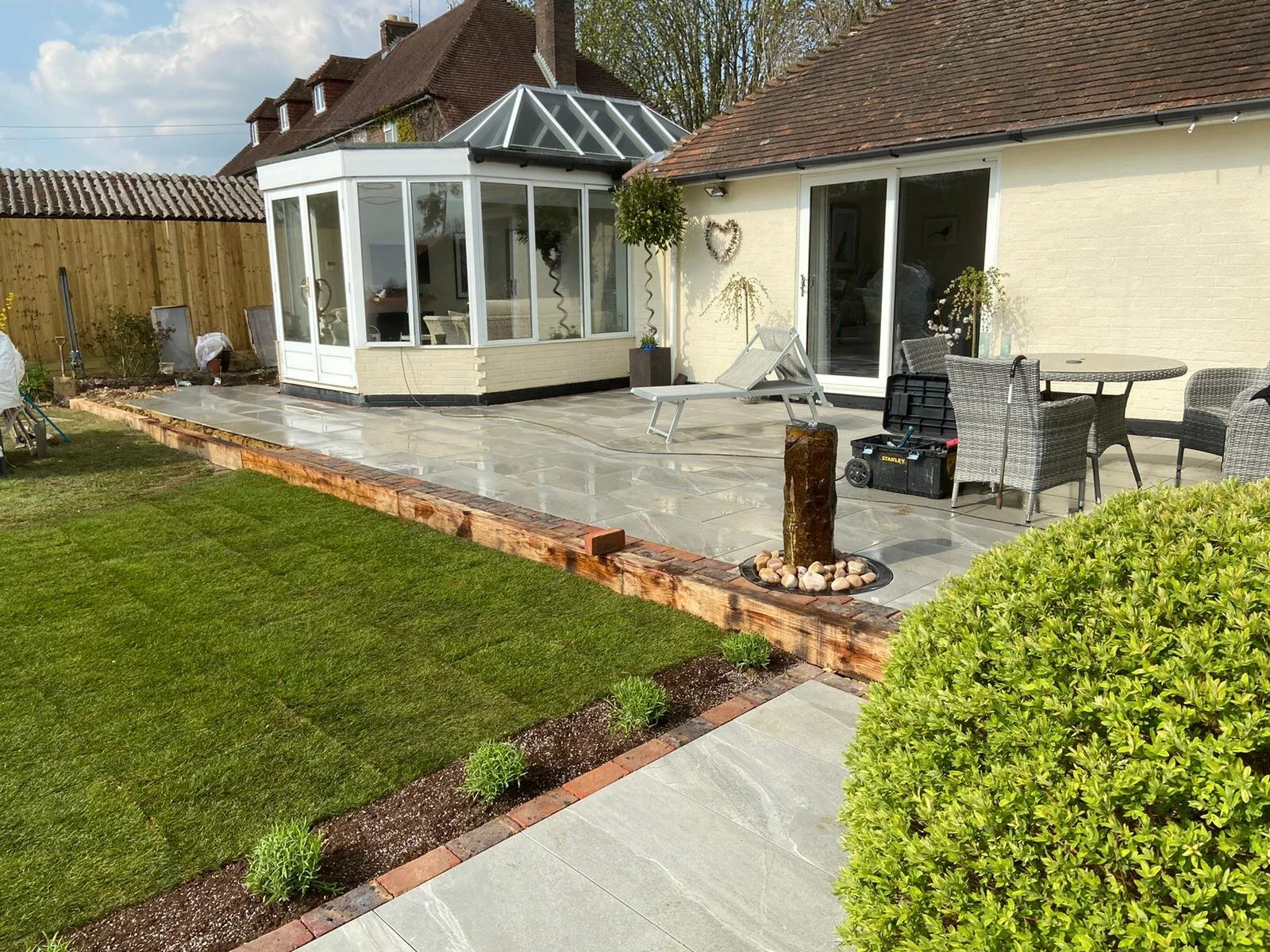 A backyard patio with newly installed stone tiles, a small water feature, outdoor furniture, and a well-maintained lawn with a flower bed.