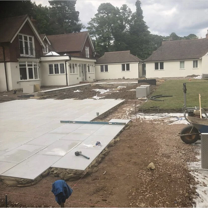 Backyard under construction with paving stones being laid, a wheelbarrow, and construction tools near a white house with multiple windows and a small porch, surrounded by trees.