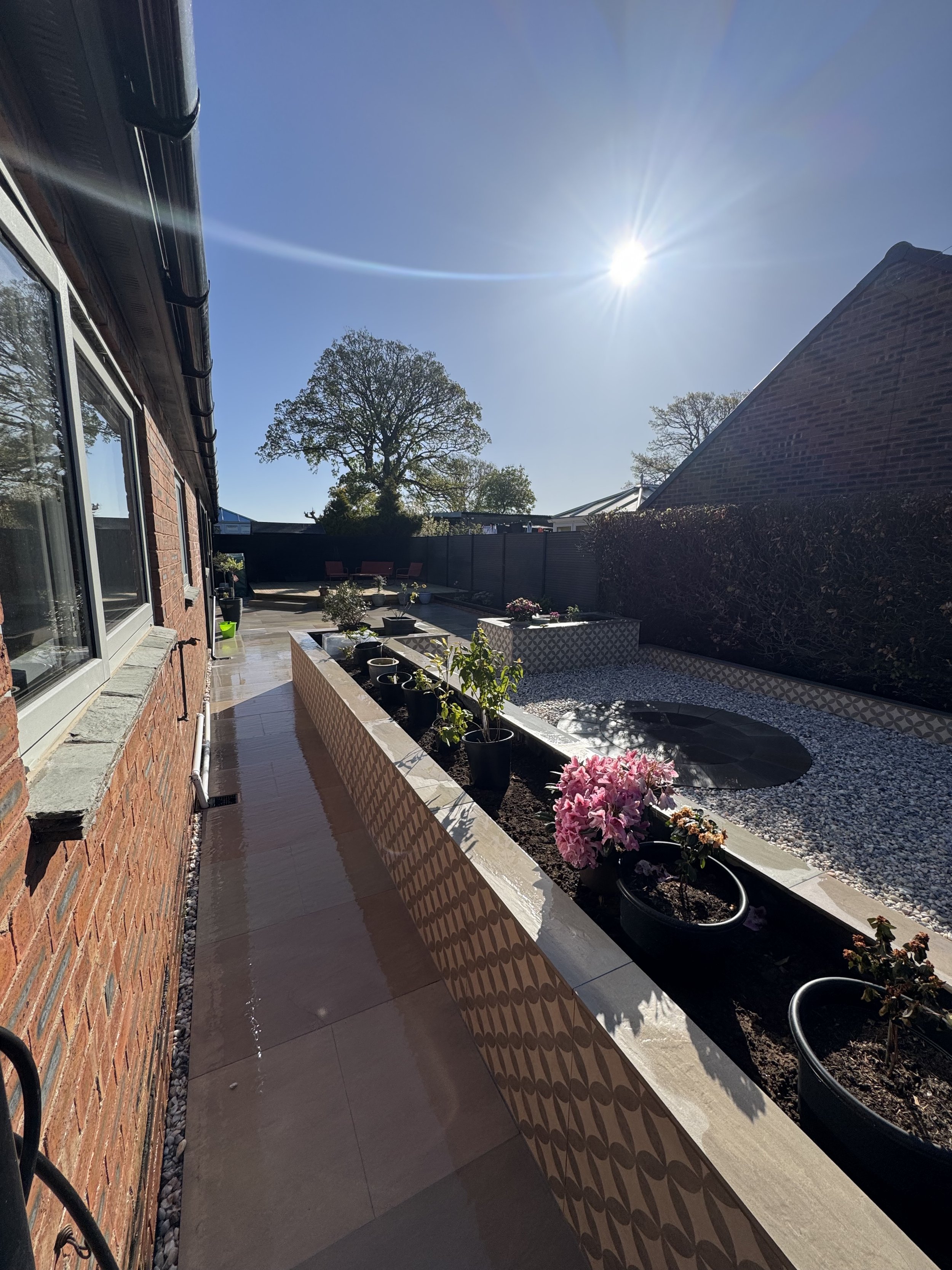 Backyard patio with potted plants, flower bed, and outdoor seating under a sunny sky.