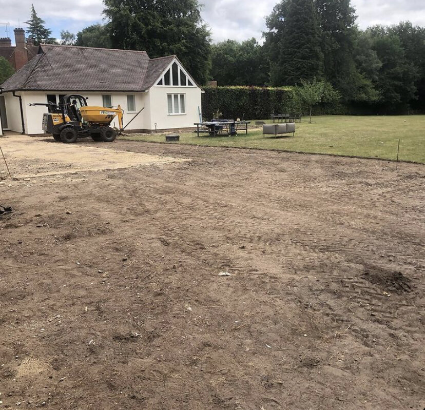 A residential backyard undergoing landscaping, with a large area of dirt, a small construction vehicle, and a white house with a gable roof and large windows surrounded by trees and greenery.