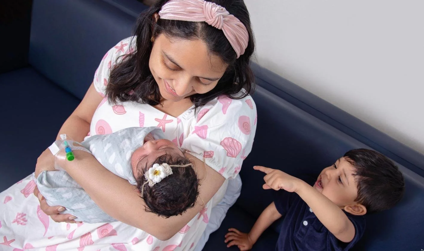 Mother cradling her newborn while big brother points during a Fresh48 hospital photo session at NewYork-Presbyterian Queens, Flushing