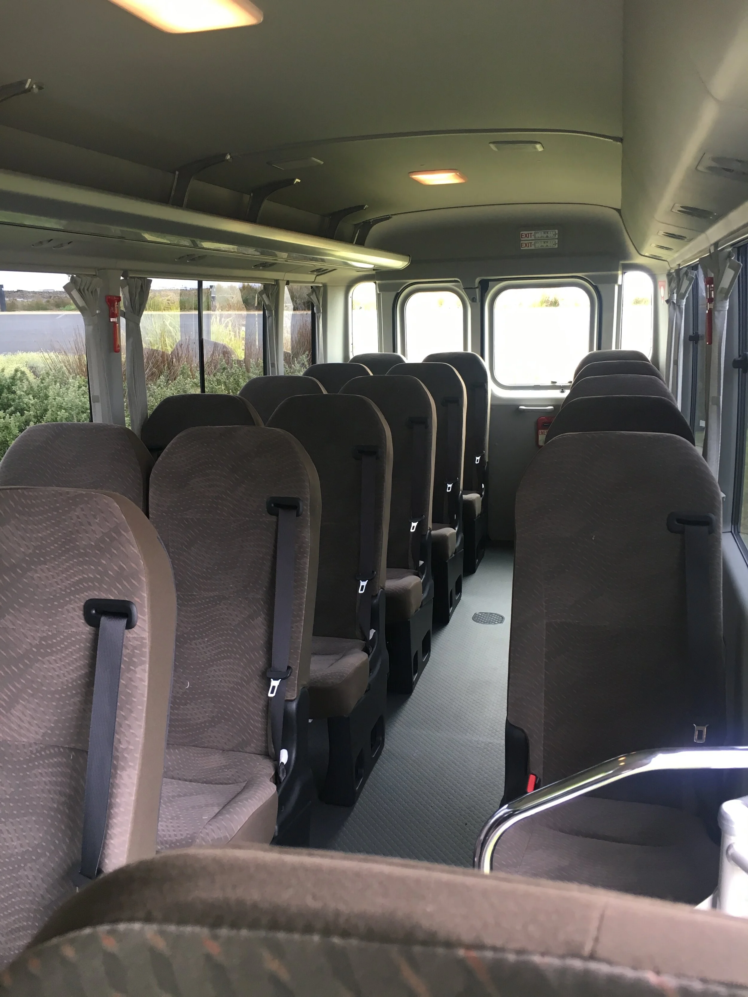 Interior of a bus with empty brown seats, windows with curtains, and an exit sign at the front.