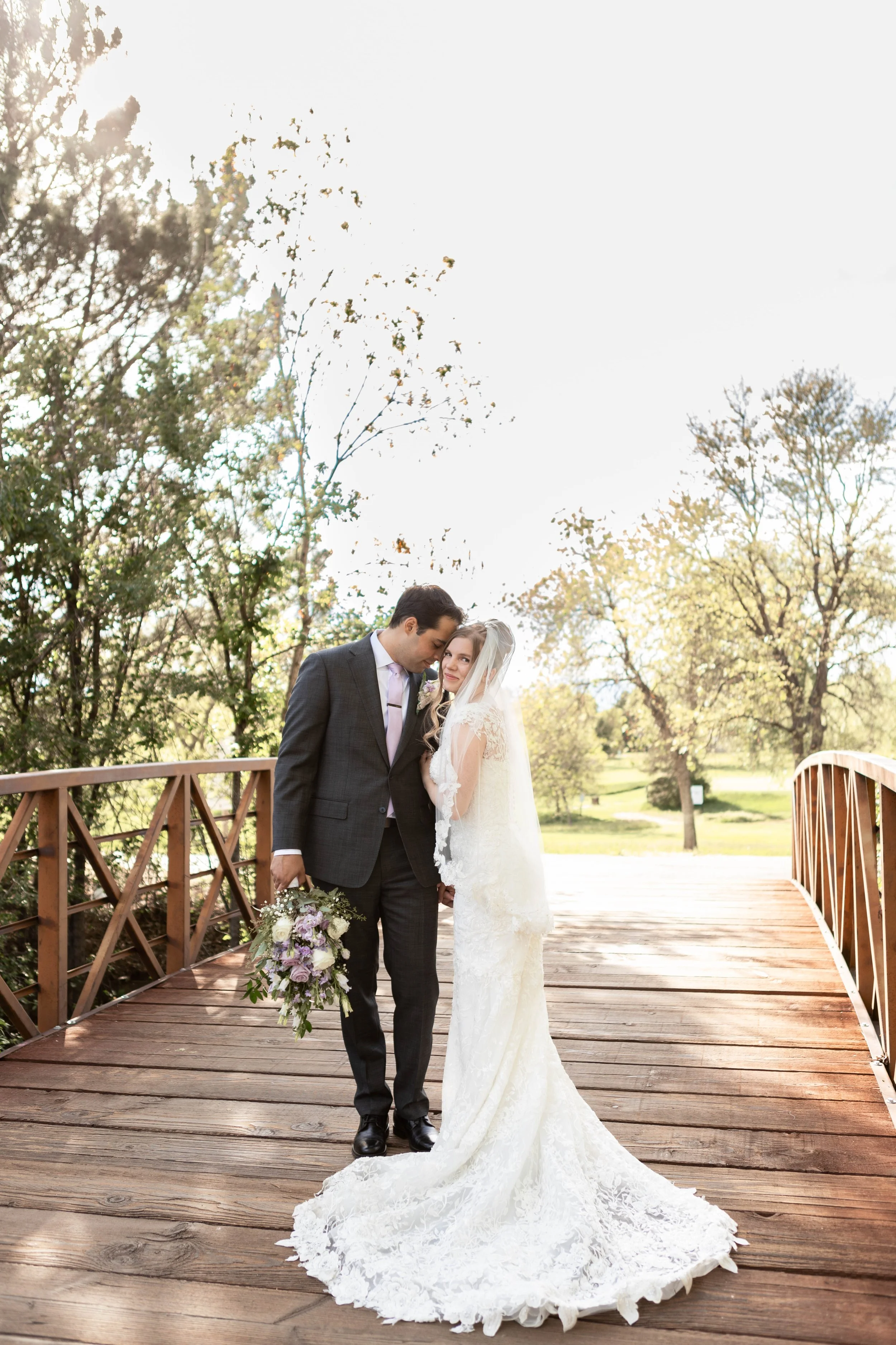 A bride and groom standing close together on a wooden bridge, surrounded by trees, with sun shining in the background, during a wedding photoshoot.