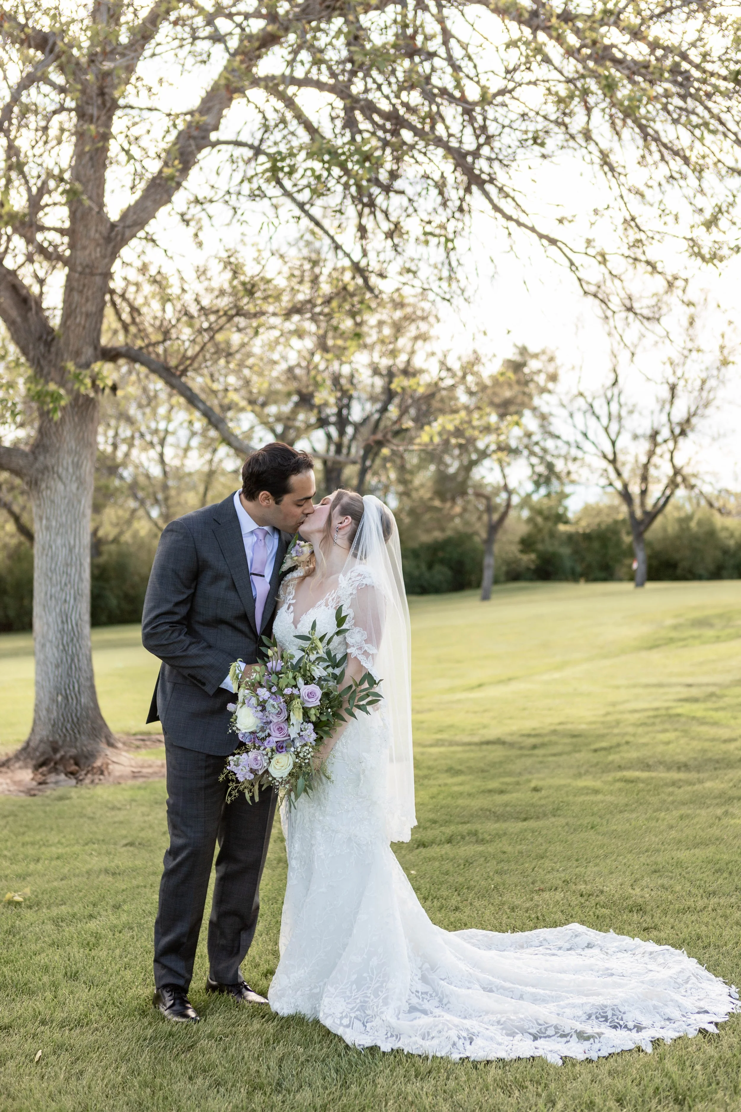 A bride and groom sharing a kiss outdoors on a grassy area with trees in the background, the bride holding a large bouquet of purple, white, and green flowers.