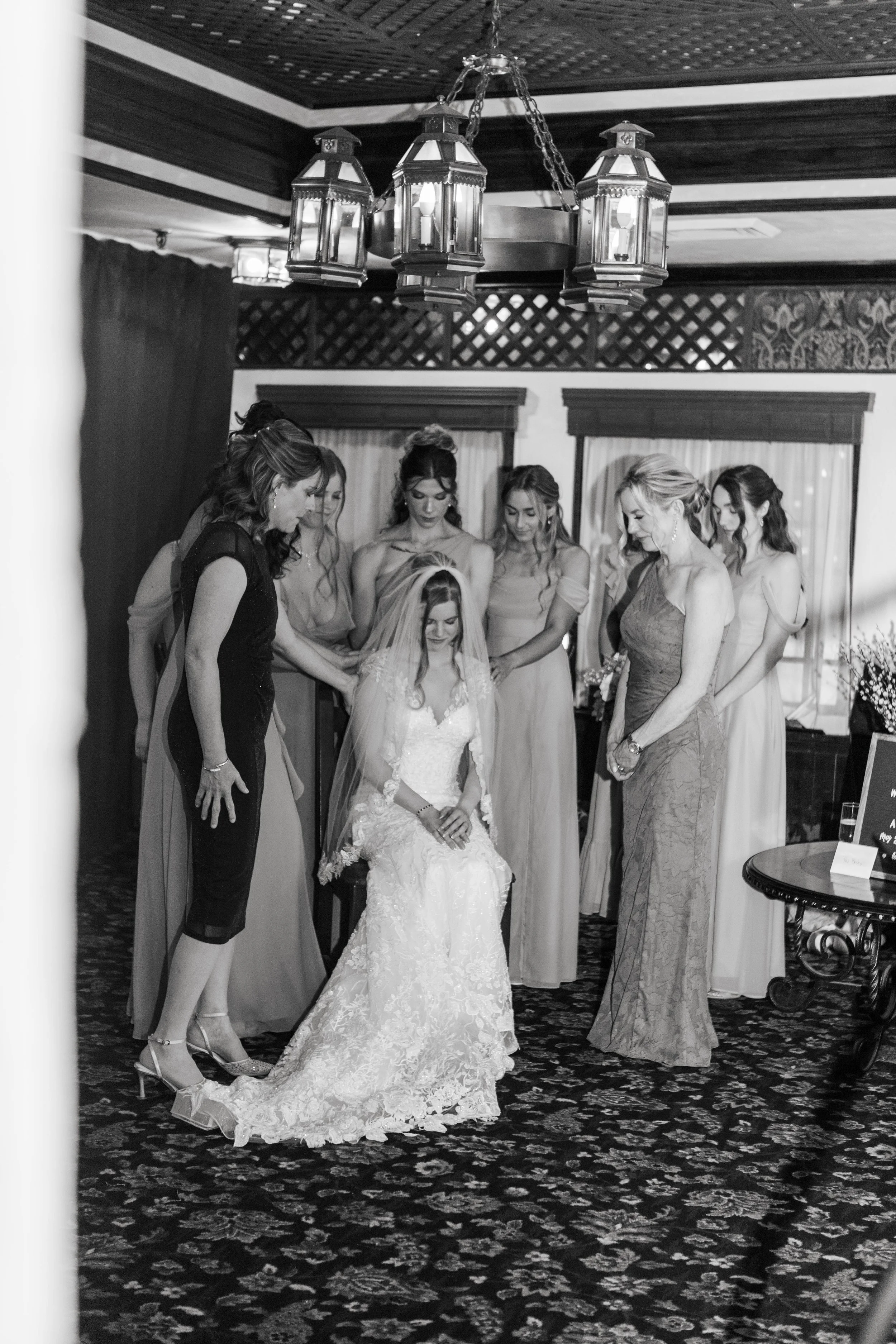 A bride in a wedding dress sitting on a chair, surrounded by six women, likely her bridesmaids, in a room with ornate ceiling details, draped curtains, and a chandelier hanging from the ceiling.