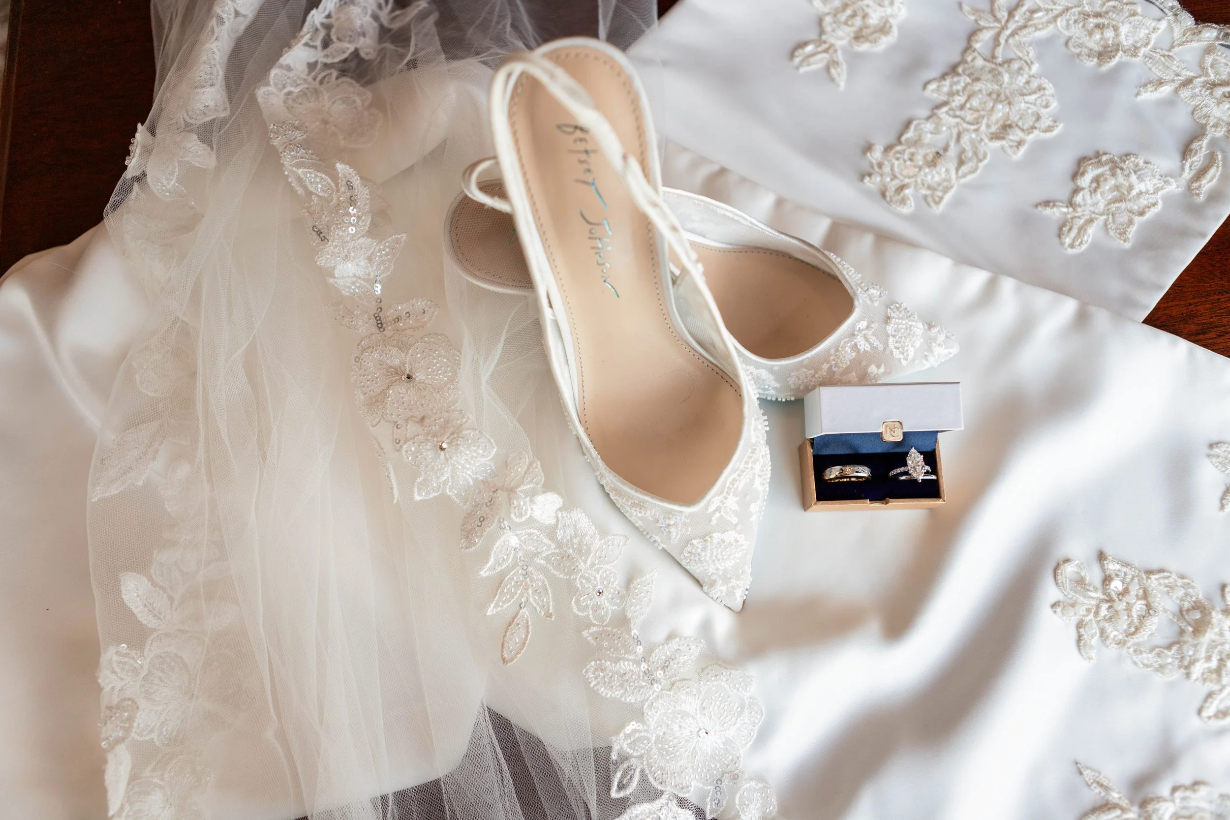 A pair of white lace high-heeled shoes, a box with rings, and a wedding veil with lace and embroidery on a satin fabric.
