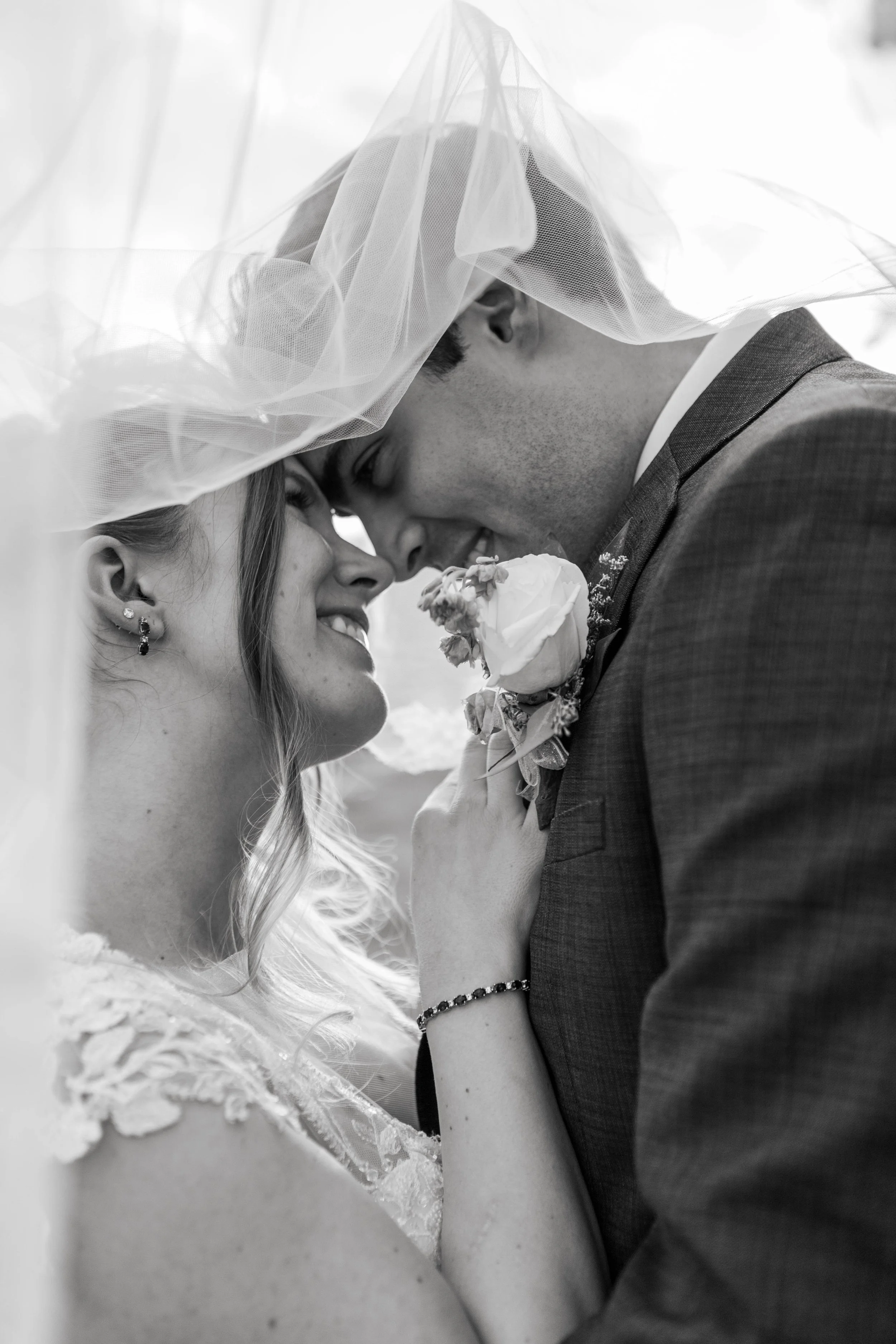 Black and white photo of a bride and groom touching heads and smiling, with a veil draped over the bride's head.