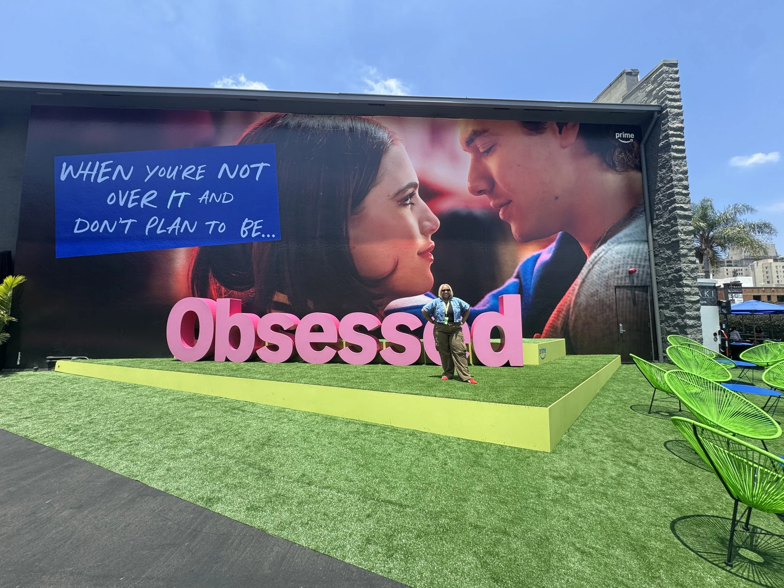 A large outdoor billboard displaying a close-up photo of a young couple facing each other, with the phrase 'When you're not over it and don't plan to be...' written on the left side. There's a woman standing in front of the billboard, smiling, and pi