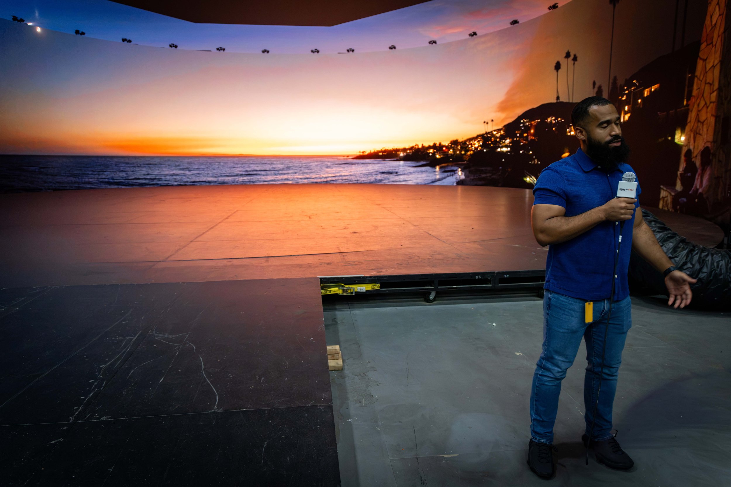 Man with beard holding a microphone in front of a large sunset projection of a beach scene with palm trees. The projection depicts a sunset over the ocean with a colorful sky and silhouette of hills and trees.