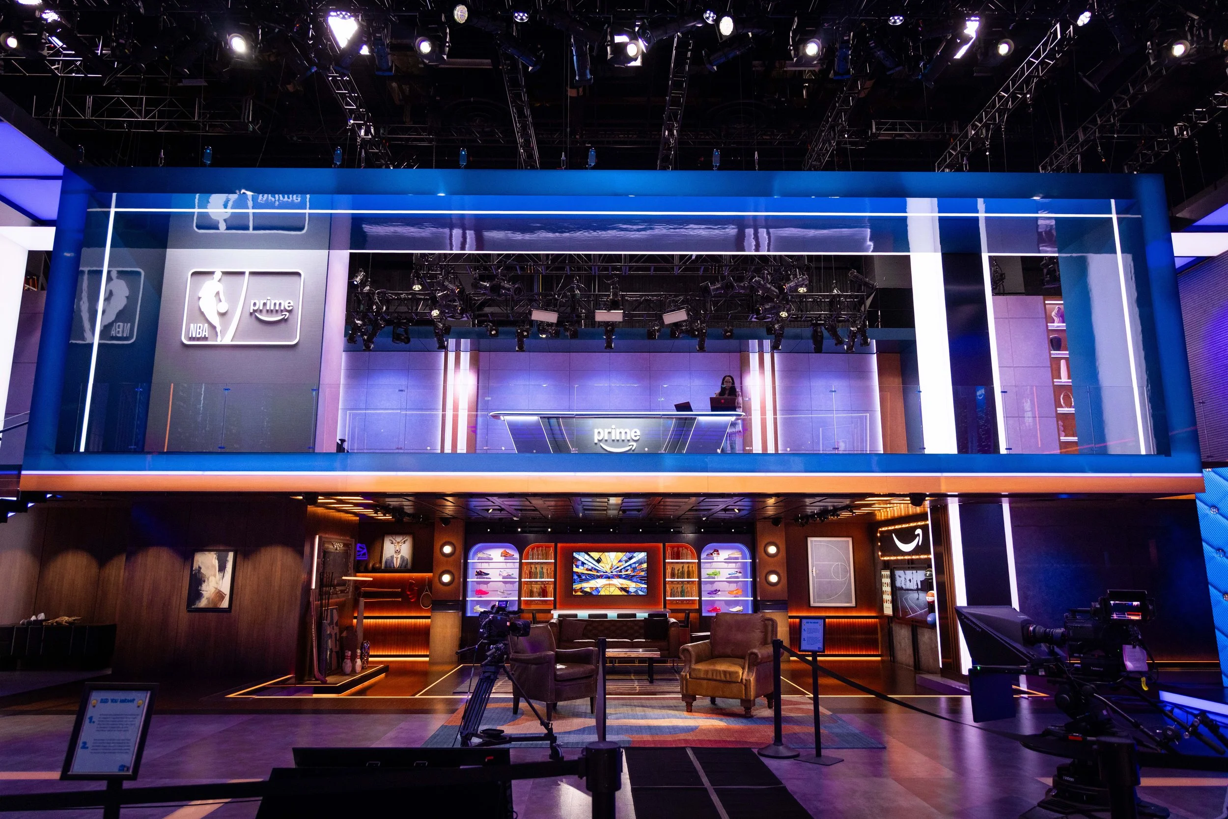 Inside view of a modern television studio or broadcasting set with blue and purple lighting, featuring a large screen displaying the 'NBA Prime' logo, and seating area with chairs and couches.