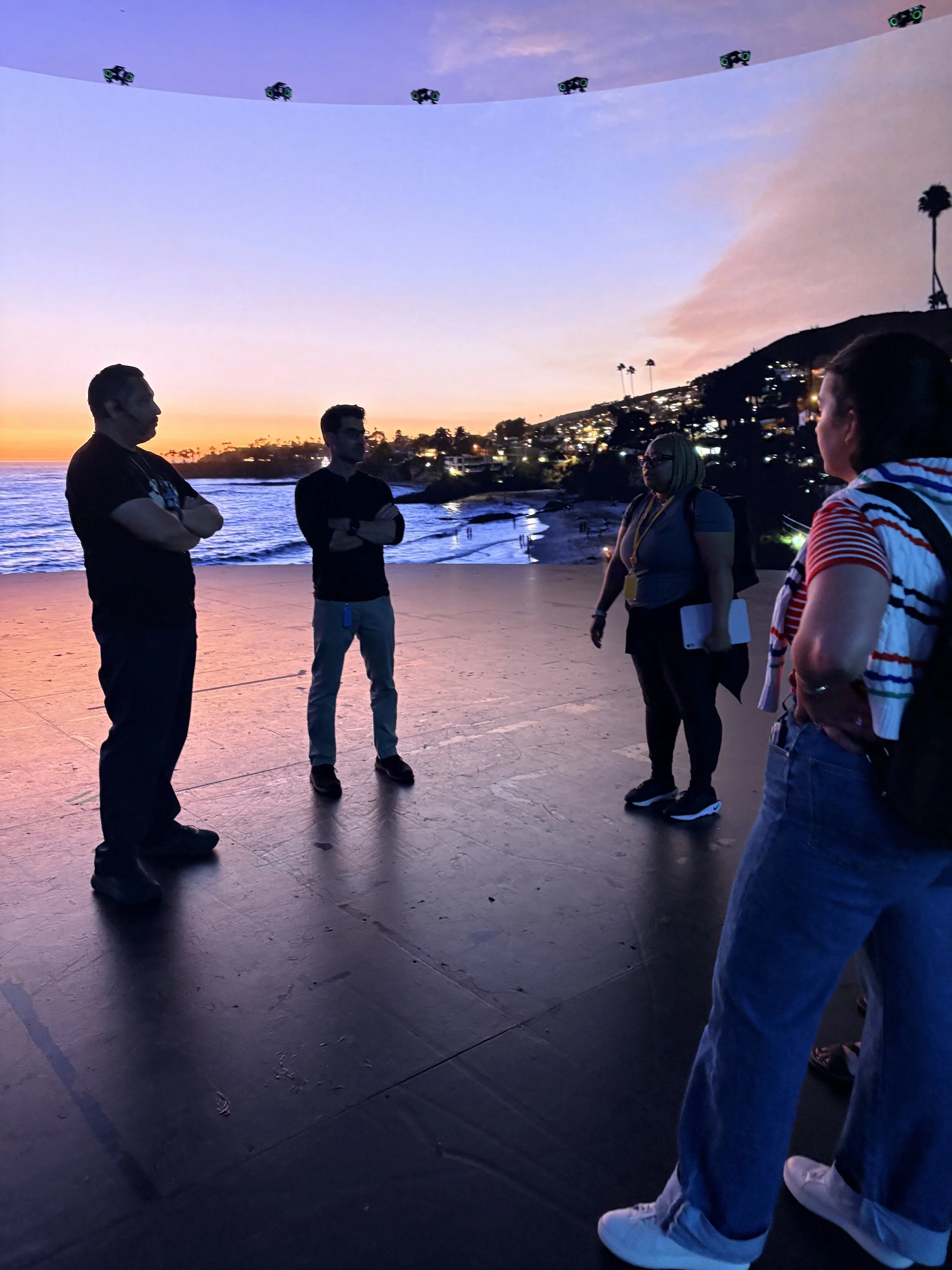A group of people standing on a paved area at sunset near the ocean, with a hill and illuminated buildings in the background. Some have sunglasses and are wearing casual clothes, engaging in conversation.