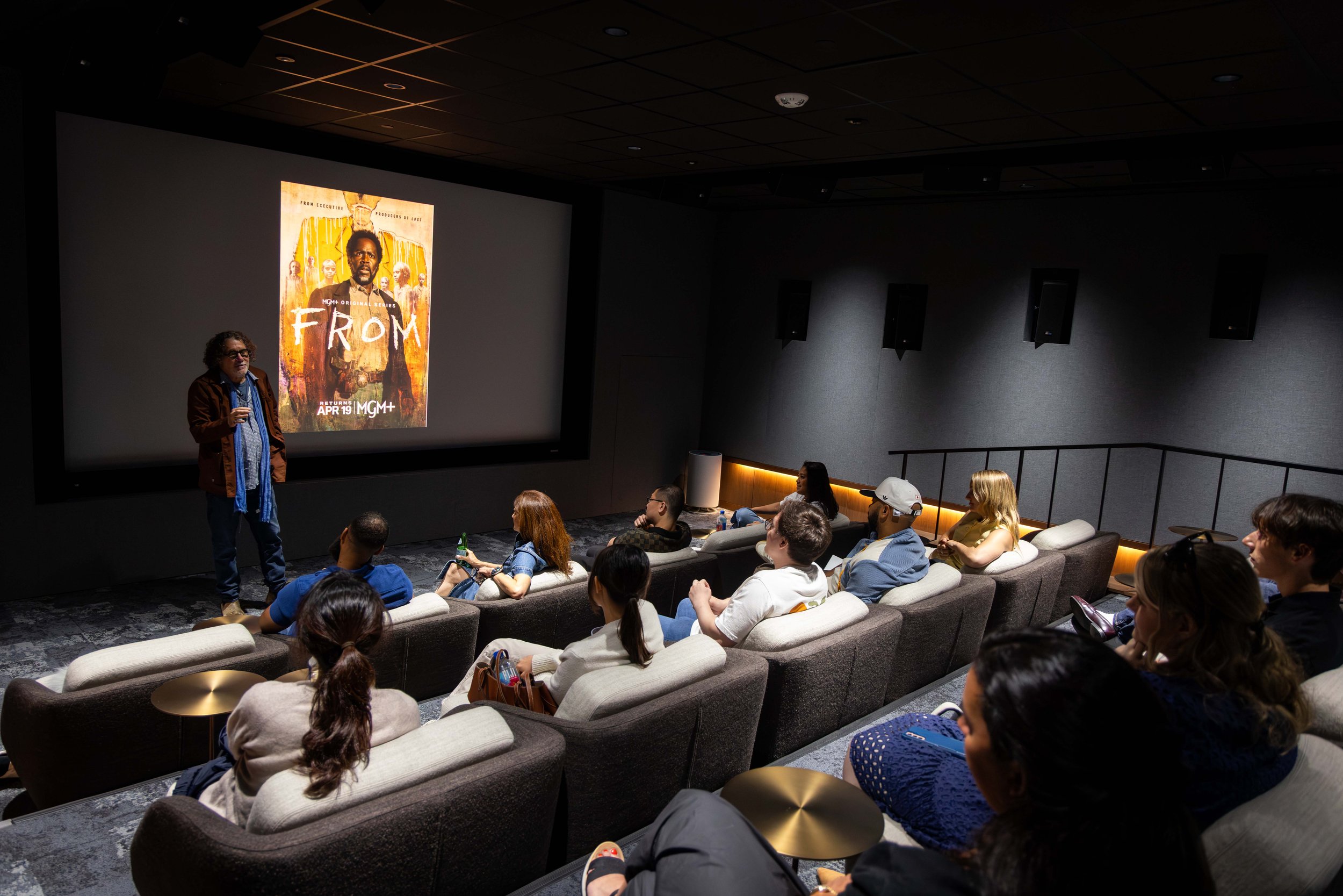 A movie theater with a group of people seated on sofas watching a presentation. A man is standing in front of a large screen displaying a movie poster for 'From.'