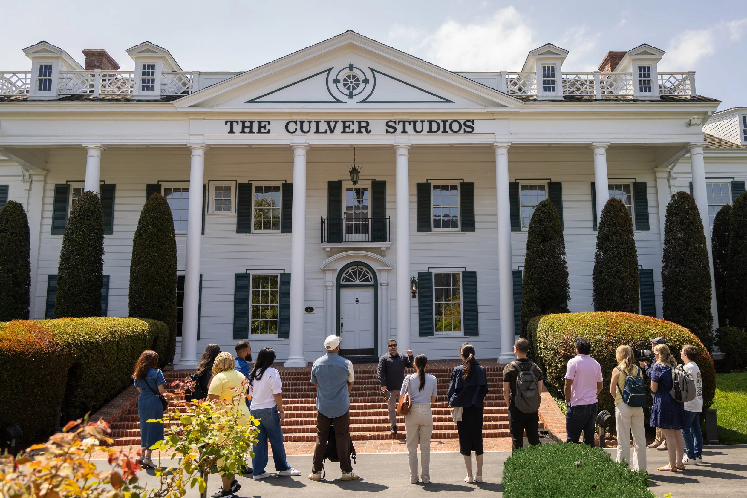 Group of people gathered outside the white colonial-style building named The Culver Studios, with brick steps, green bushes, and trees.