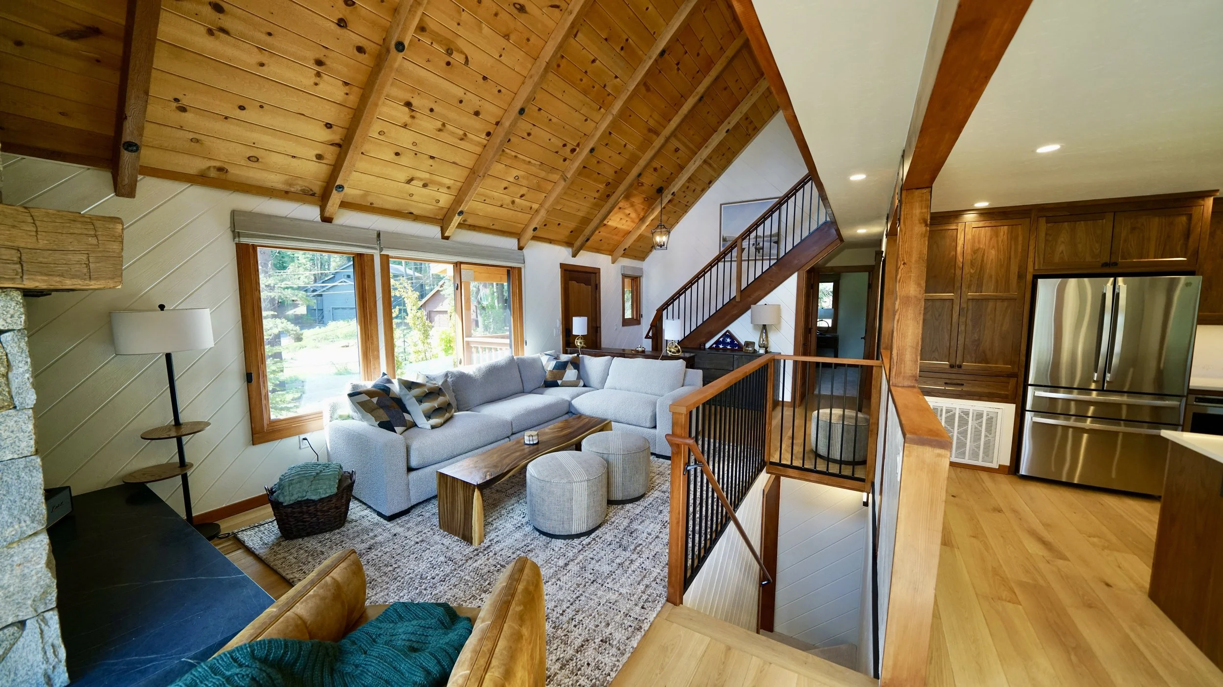 Living room with a vaulted wooden ceiling, large window, white sectional sofa, wooden coffee table, and two round ottomans. Kitchen with wooden cabinets and stainless steel refrigerator is visible in the background.