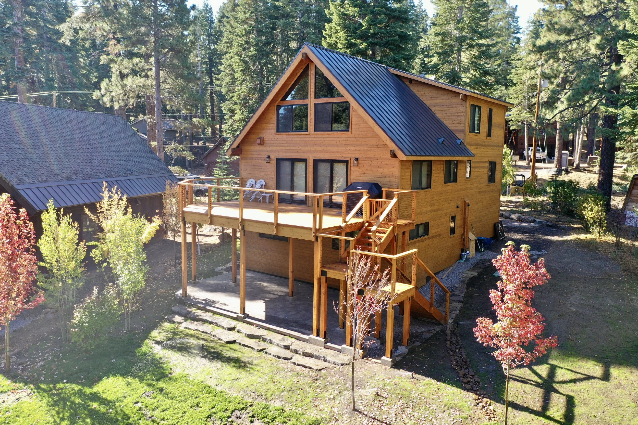 A wooden house with a blue metal roof in a forested area. It has a deck with outdoor chairs and a grill, surrounded by trees with autumn leaves.