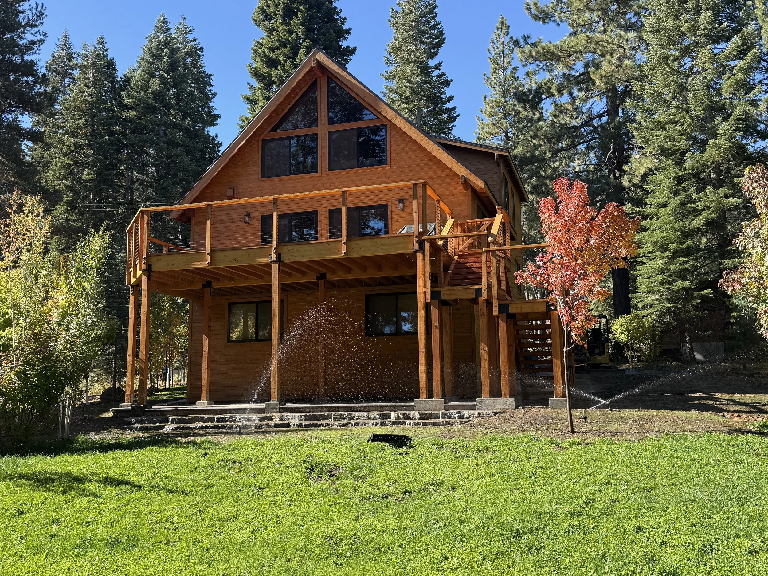 A wooden house with a steep roof and large windows in a forest setting, with a green lawn, trees, and a sprinkler watering the grass.