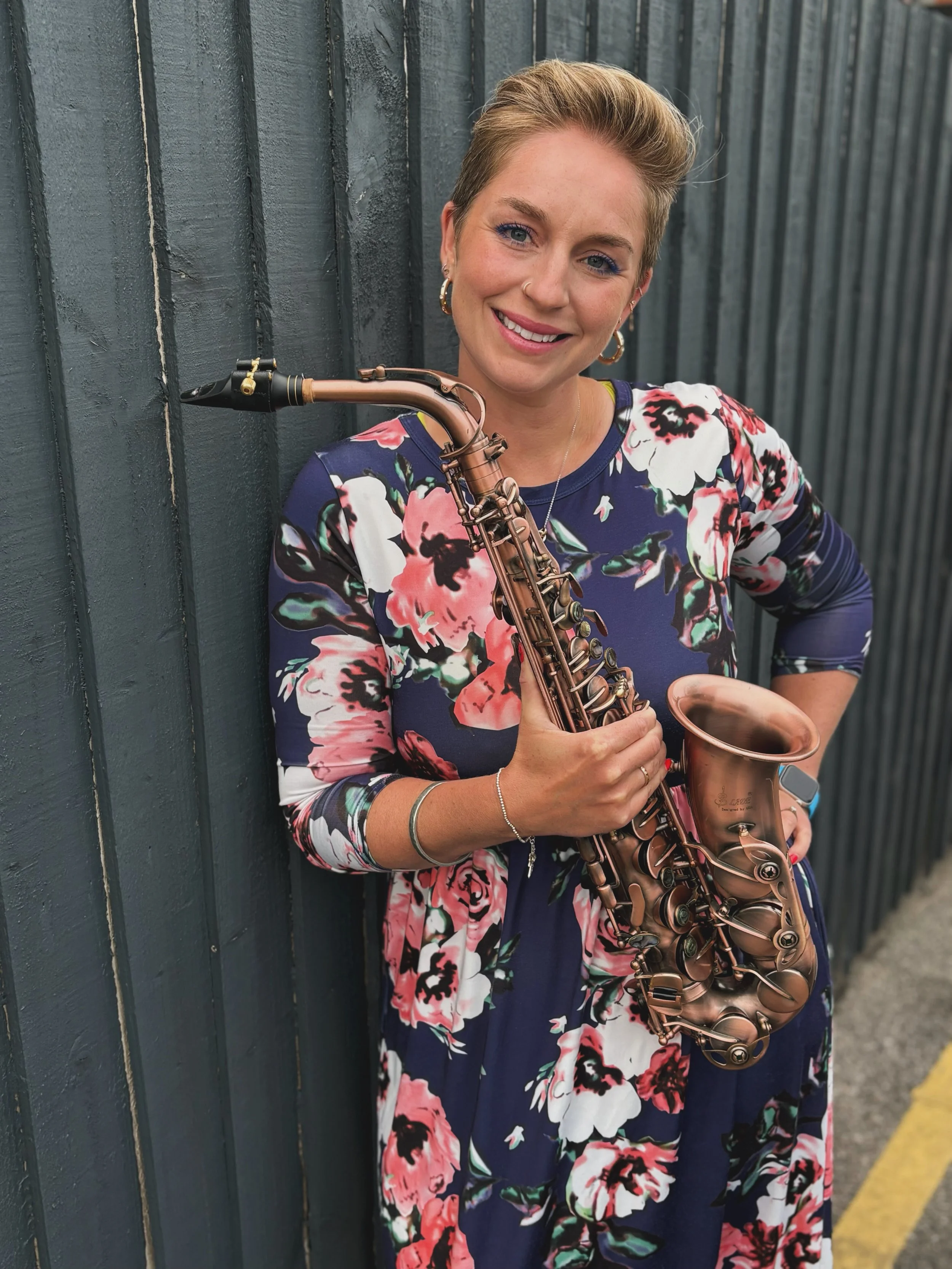 A woman with short blond hair holding a saxophone and smiling, standing against a dark-colored wooden wall, wearing a floral dress.