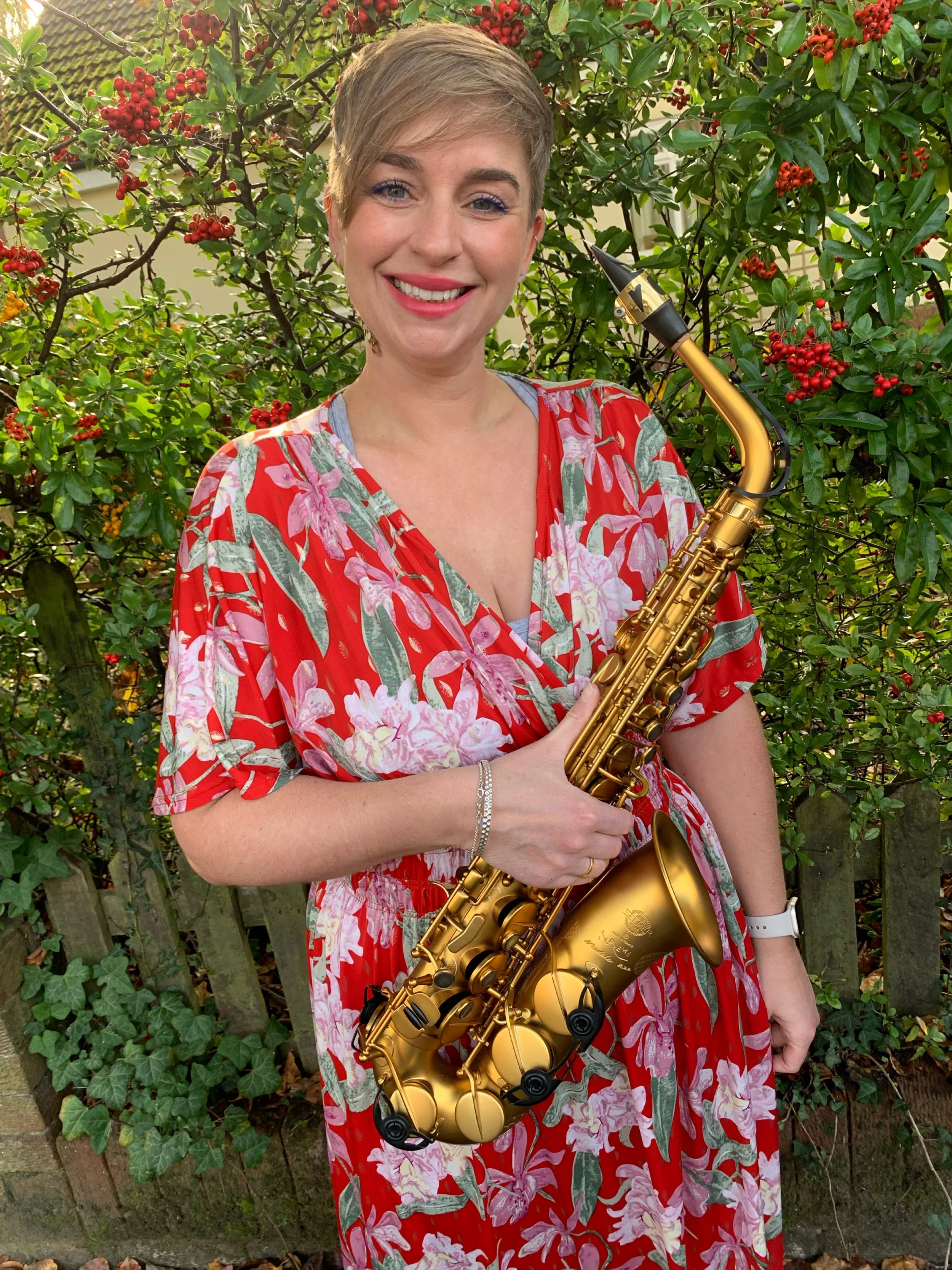 A smiling woman with short blonde hair wearing a red floral dress holding a gold saxophone while standing outdoors in front of green bushes with red berries.