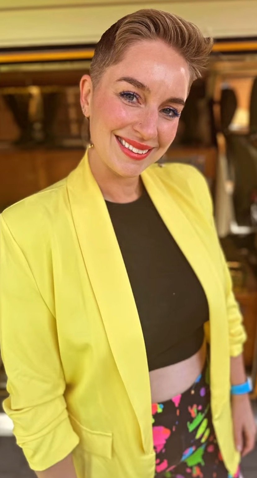 A smiling woman with short hair wearing a yellow blazer over a black crop top and colorful patterned pants, standing indoors with a blurred background.