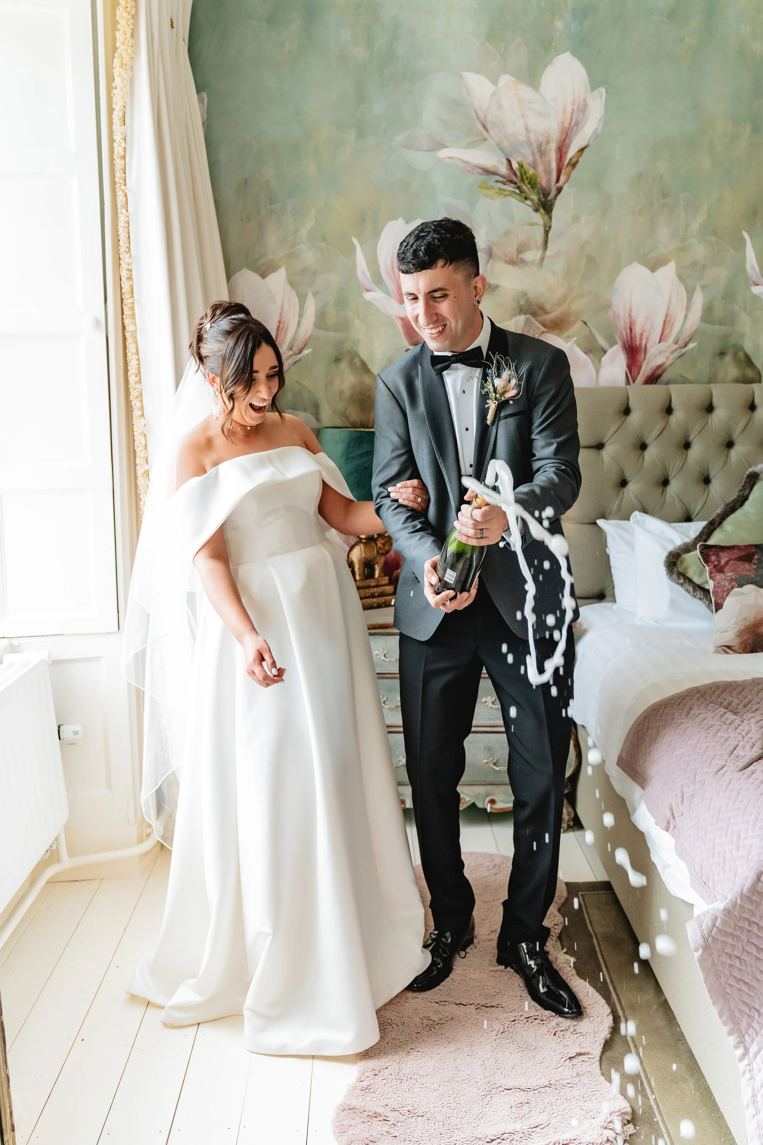 A bride and groom celebrating their wedding in a room, opening a bottle of champagne, which is spraying foam, with happy expressions.