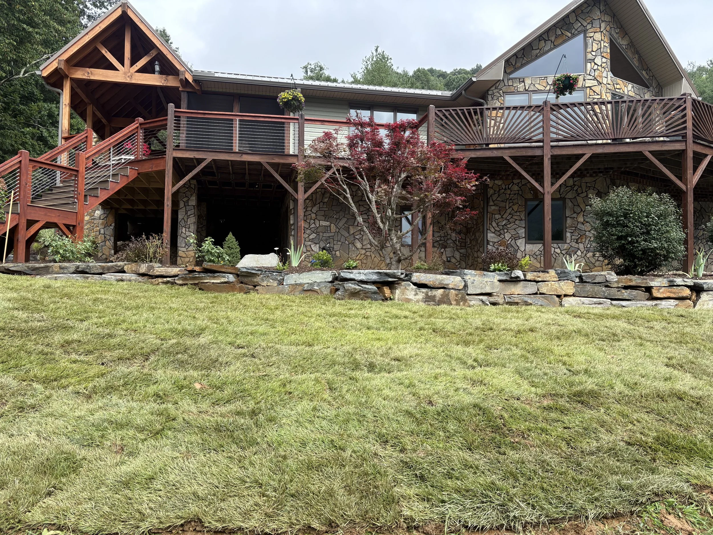 A large house with stone and wood siding, a sloped roof, and multiple decks with railings. There are trees and plants in front of the house, with a grassy lawn in the foreground.