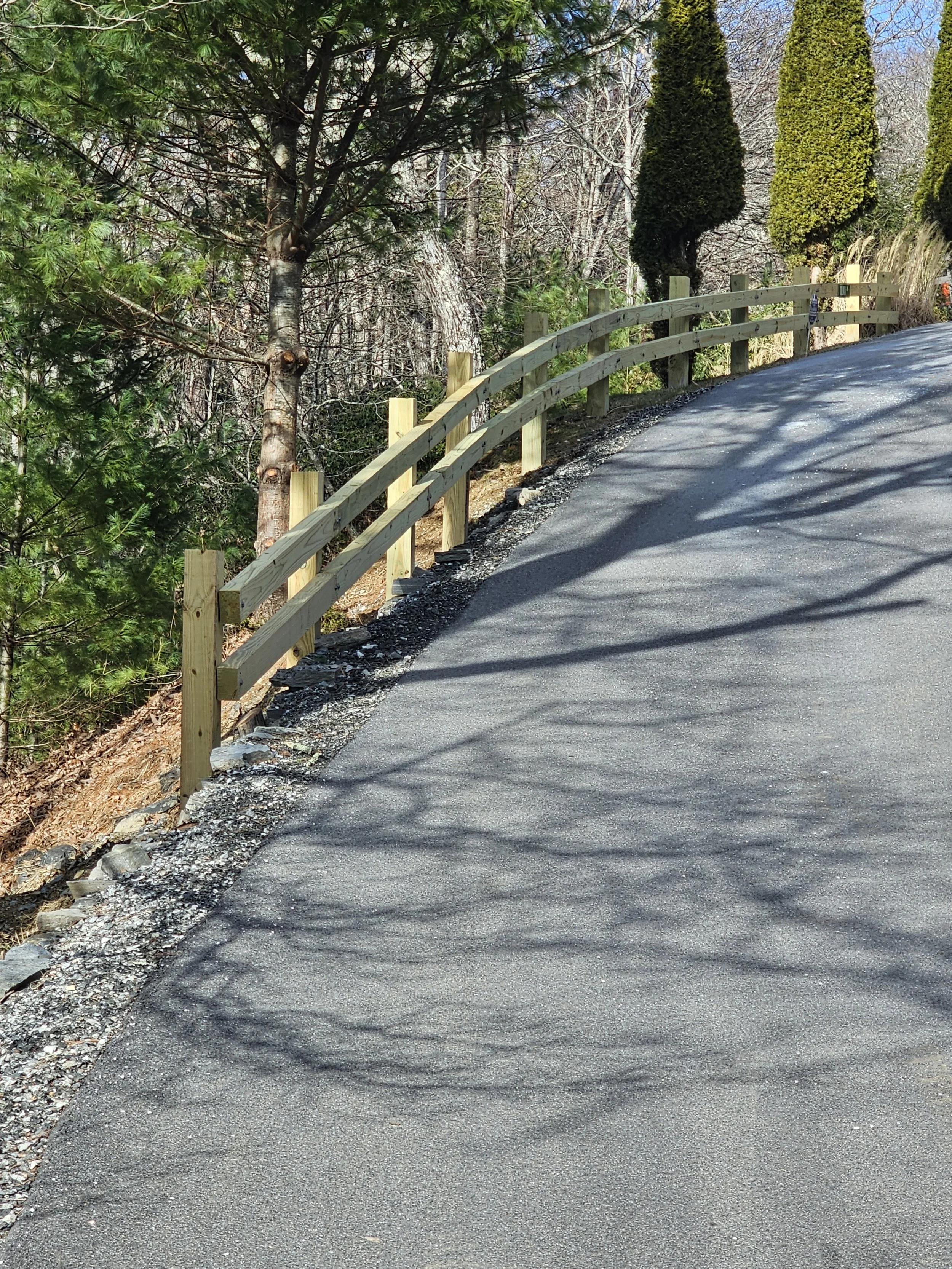 A paved road on a hillside with a wooden guardrail on the left side, trees casting shadows on the road, and tall, well-shaped evergreen bushes in the background.
