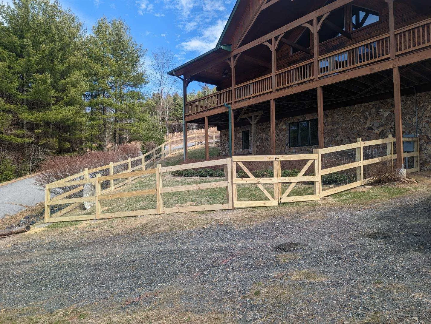 A wooden fence surrounds a gravel area beside a house with a stone and wood exterior, situated on a sloped hillside with trees and a clear blue sky in the background.