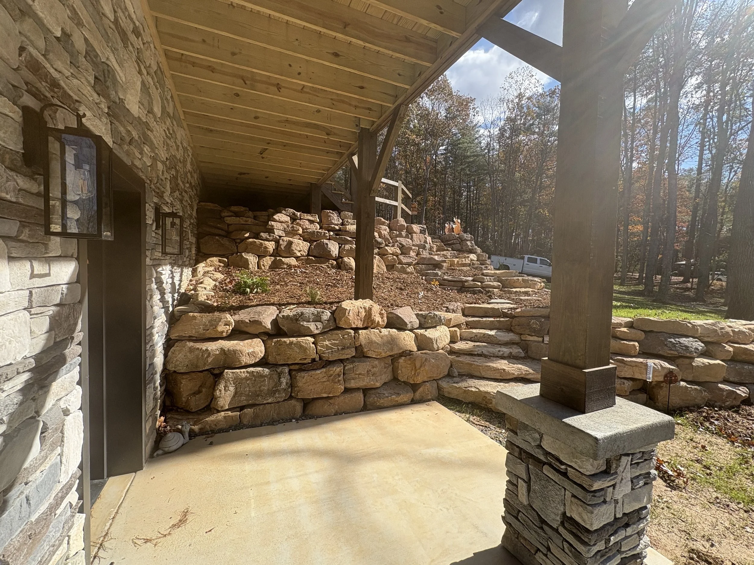View of a porch with stone walls and wooden beams, overlooking a landscaped yard with stone steps and a wooded backdrop.