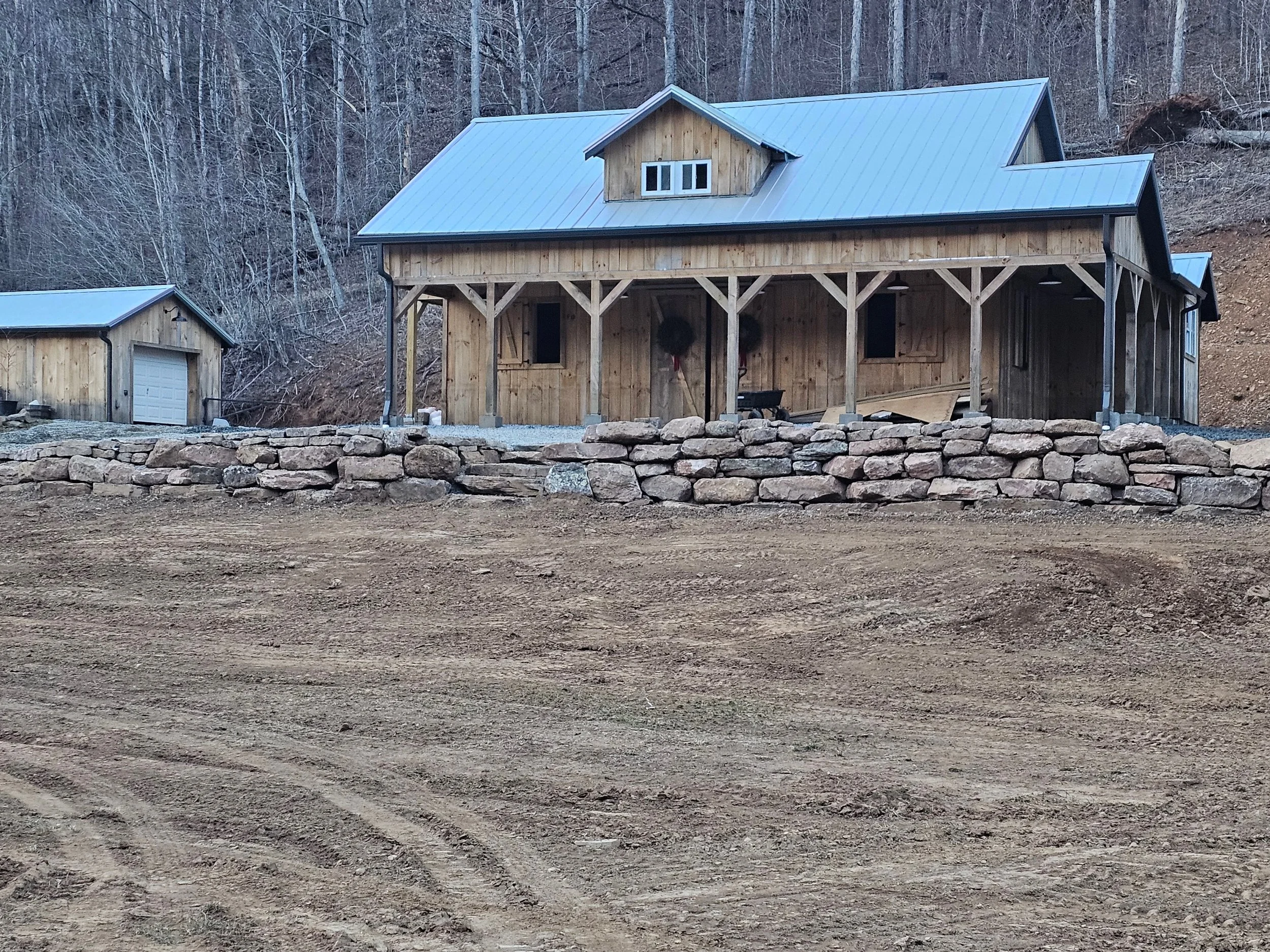 A wooden house with a metal roof on a stone foundation, surrounded by trees and a dirt area in the foreground.