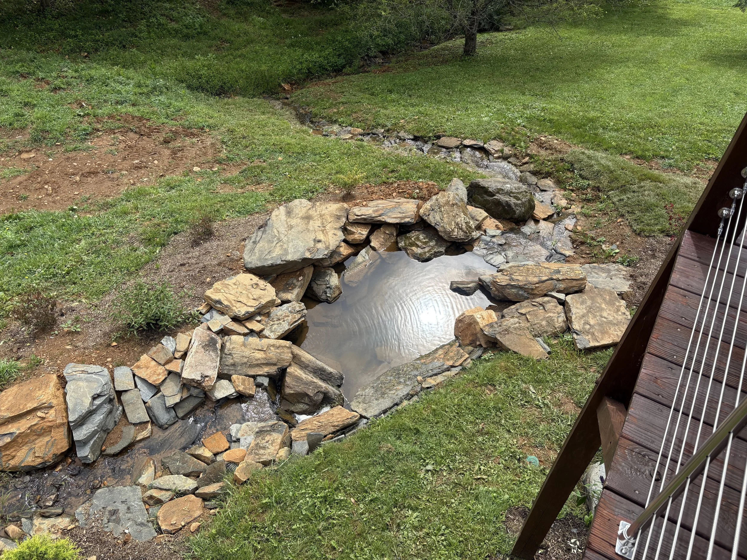 Small pond surrounded by rocks and a garden, viewed from a deck with stairs.