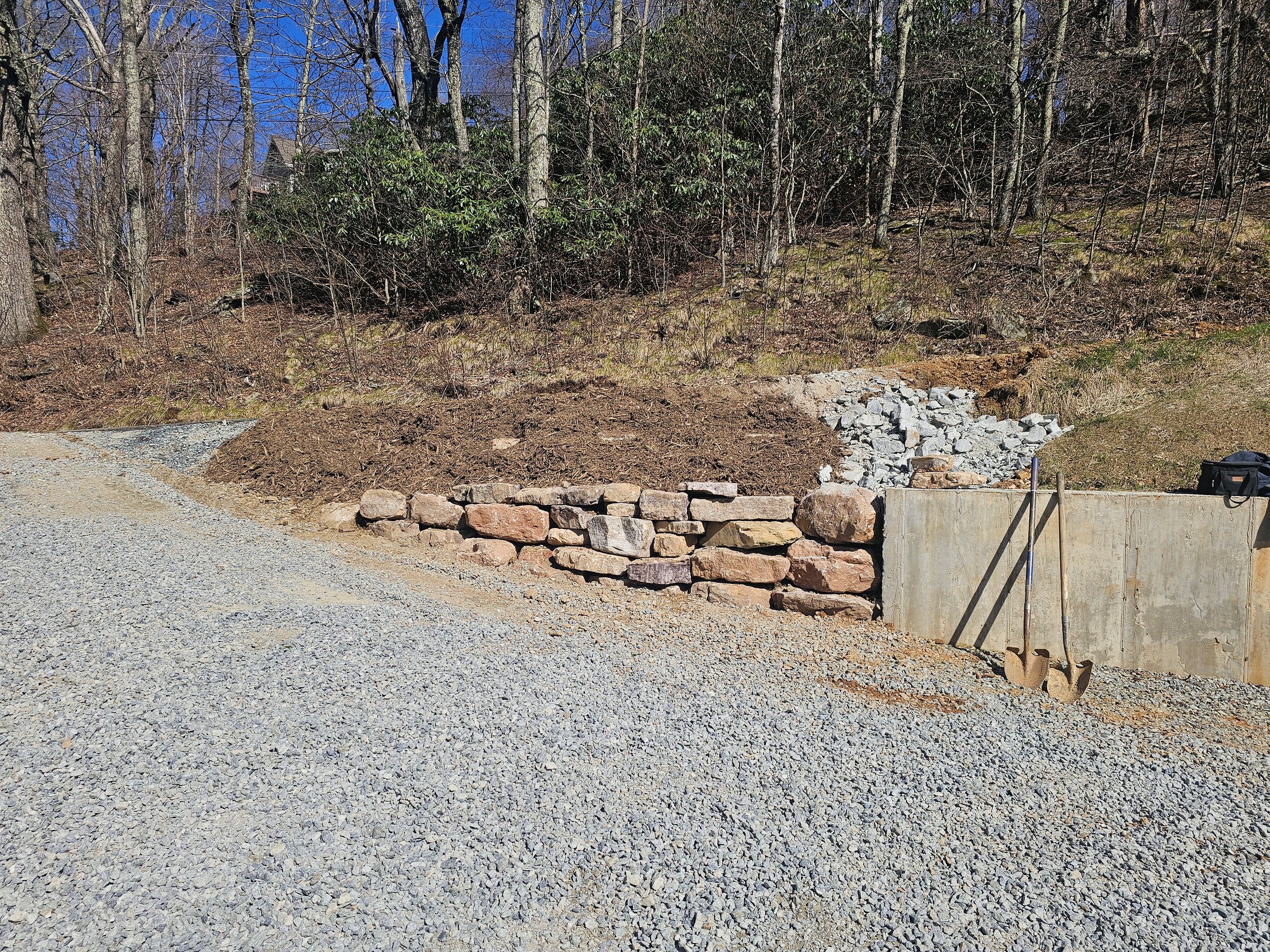 Construction site with a gravel road, a partially built stone wall, and a concrete wall with construction tools leaning against it. There are trees and a house in the background.