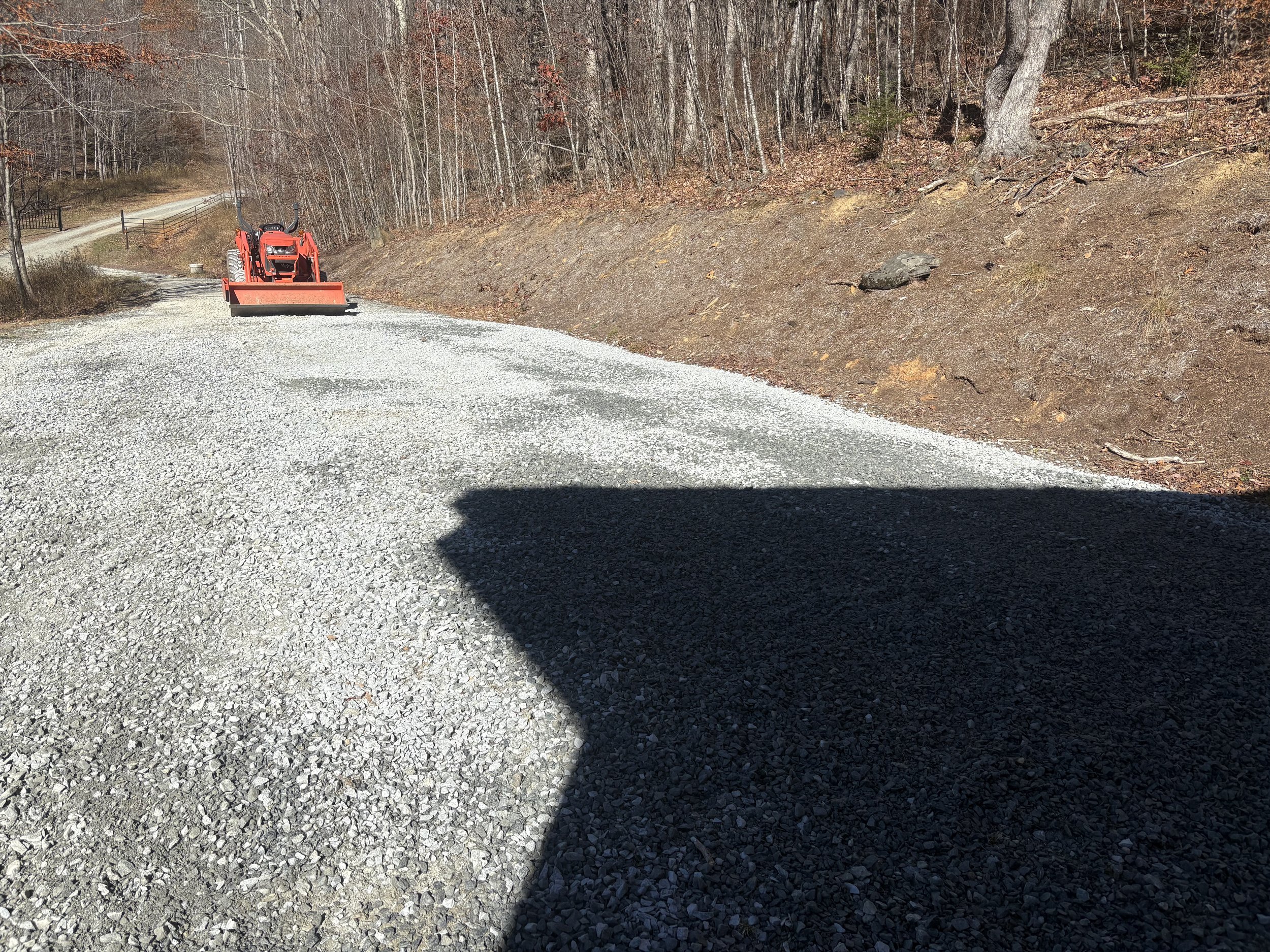 A gravel driveway on a slope with a red tractor and snow plow attachment parked at the top. The surrounding area has leafless trees and a fence, indicating a late fall or winter season.