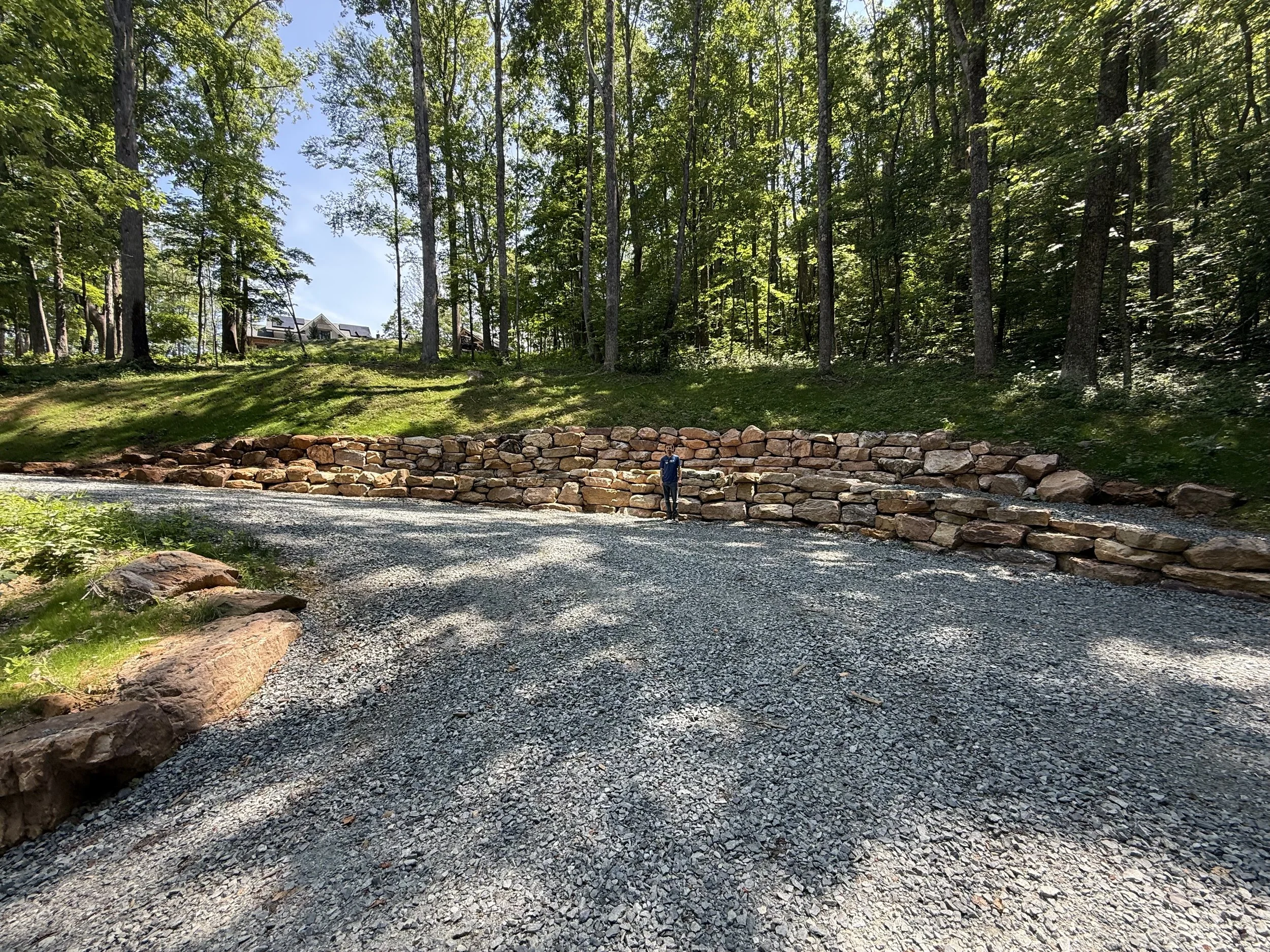 A person standing on a gravel driveway next to a stone retaining wall in a wooded area with tall trees and a house visible in the distance.