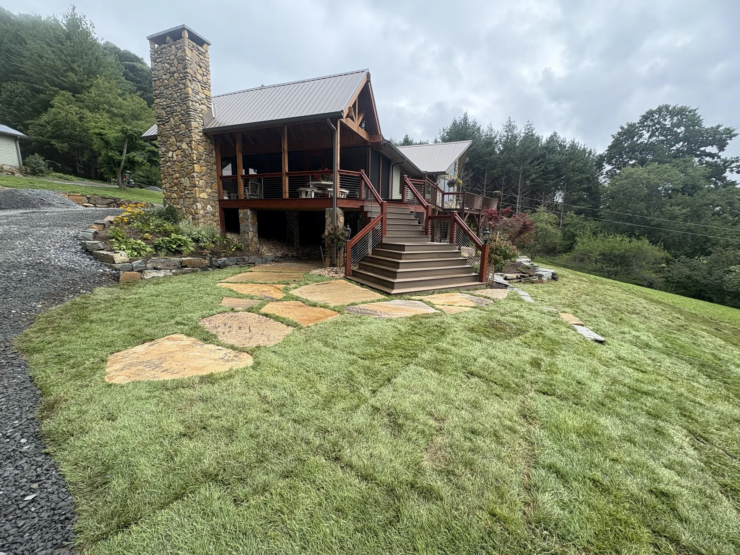 A house with a stone chimney and wooden porch, surrounded by a well-maintained lawn with stepping stones and a landscaped garden, in a wooded area under a cloudy sky.