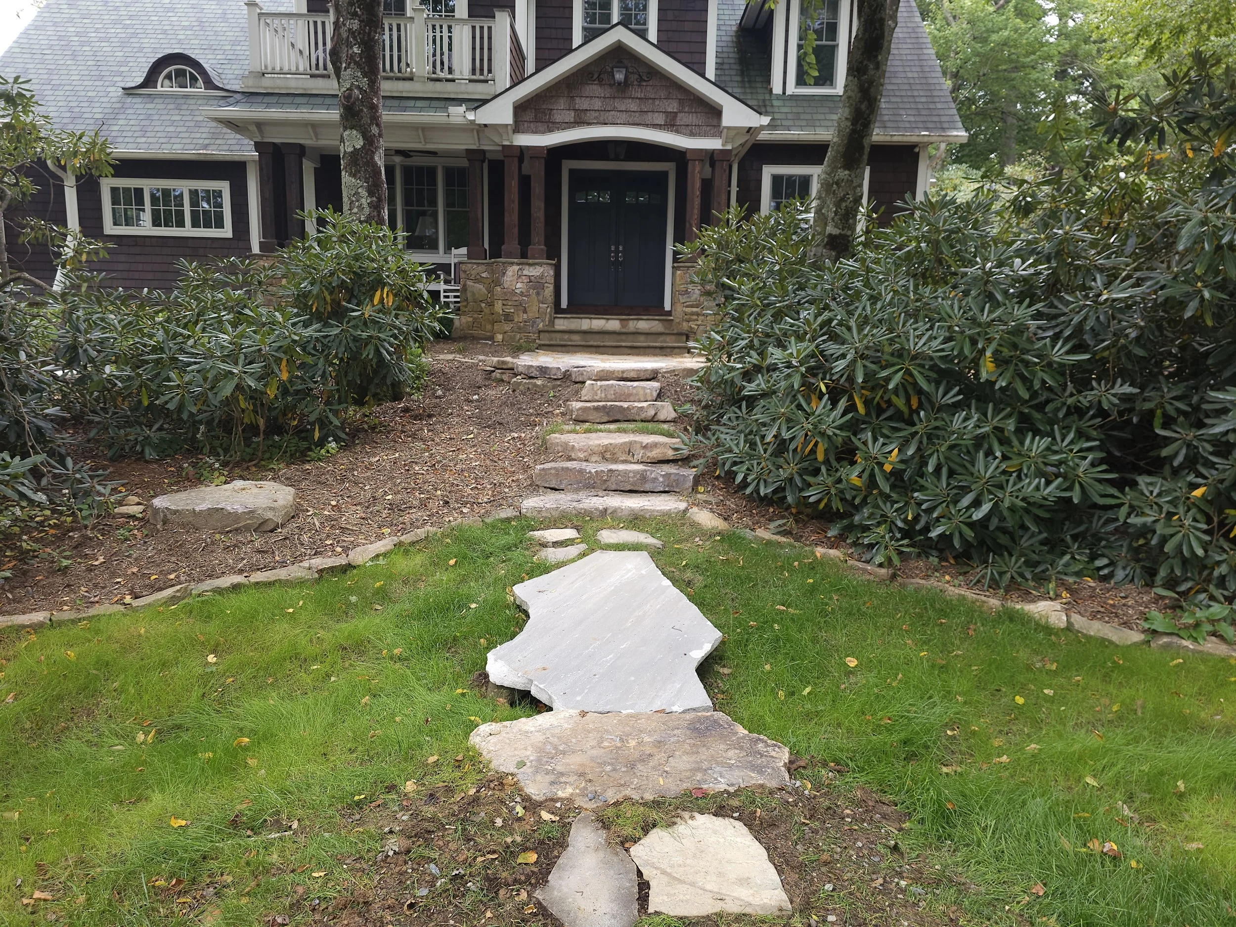 Stone pathway leading up to the front porch of a house with a dark blue door, surrounded by trees and bushes, with steps and landscaping in the front yard.