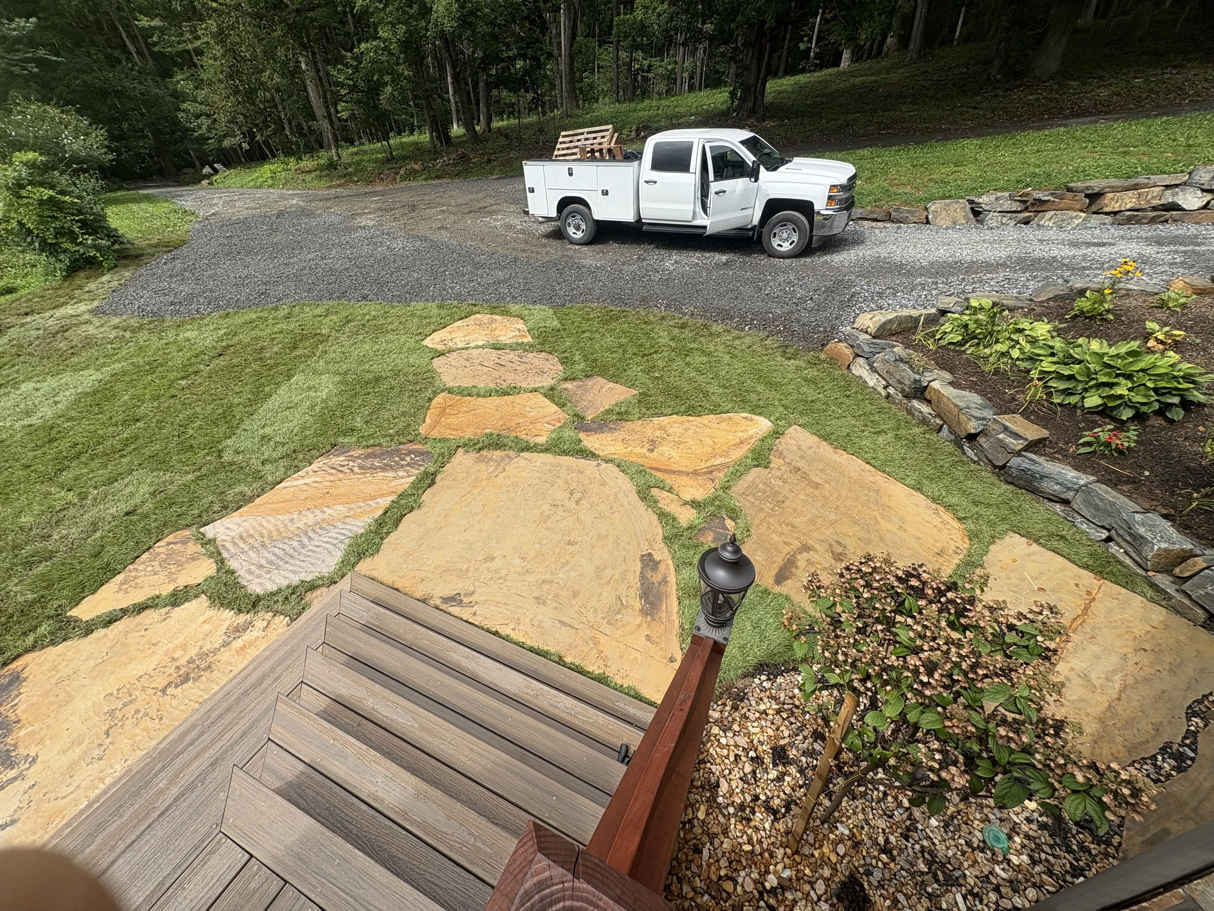 View of a front yard with a stone pathway, a small tree with pink flowers, outdoor lighting, and a wooden porch, with a gravel driveway and a white pickup truck in the background