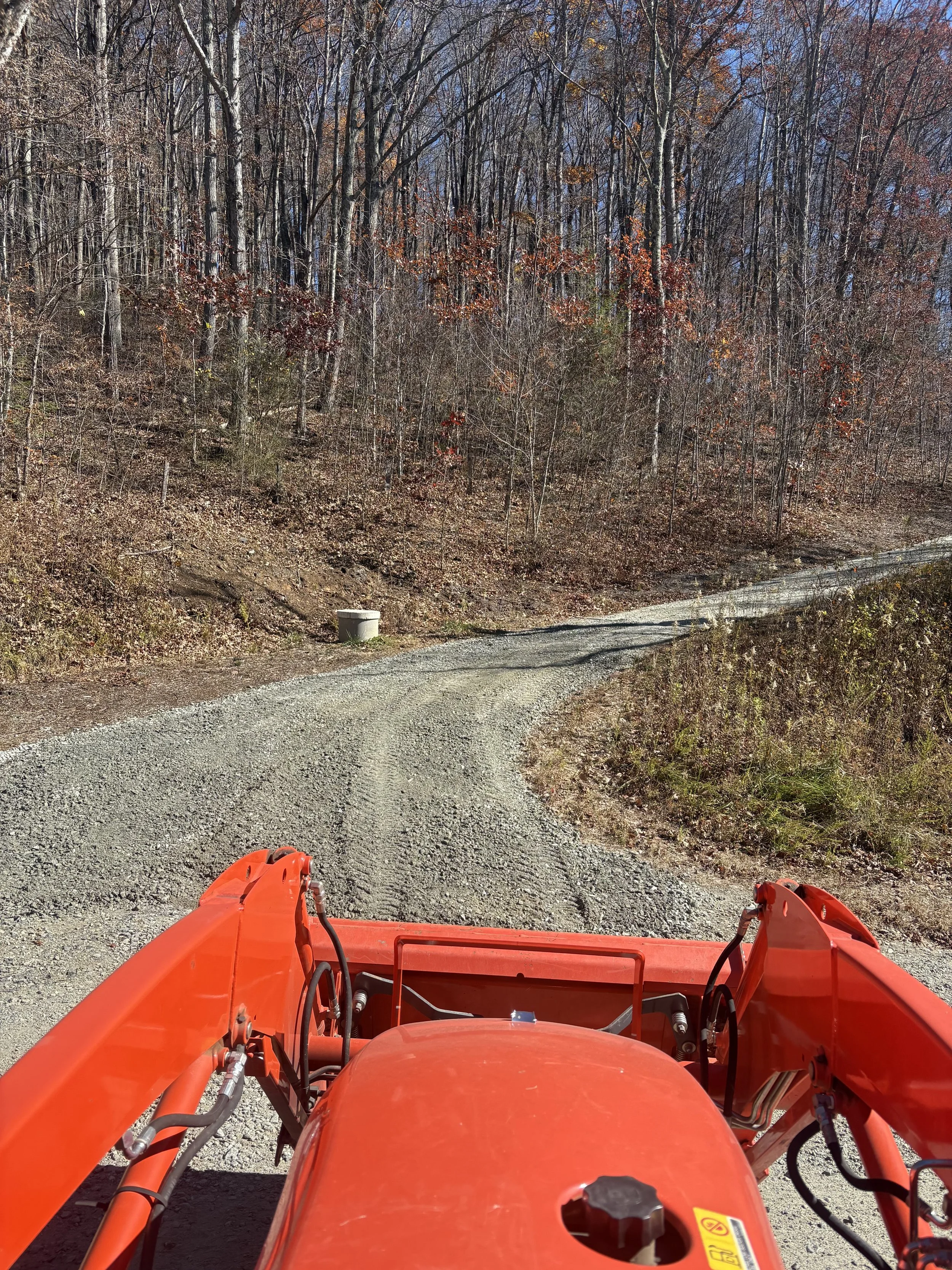 View from a red tractor on a dirt gravel trail curving through a wooded area with leafless trees and autumn foliage.