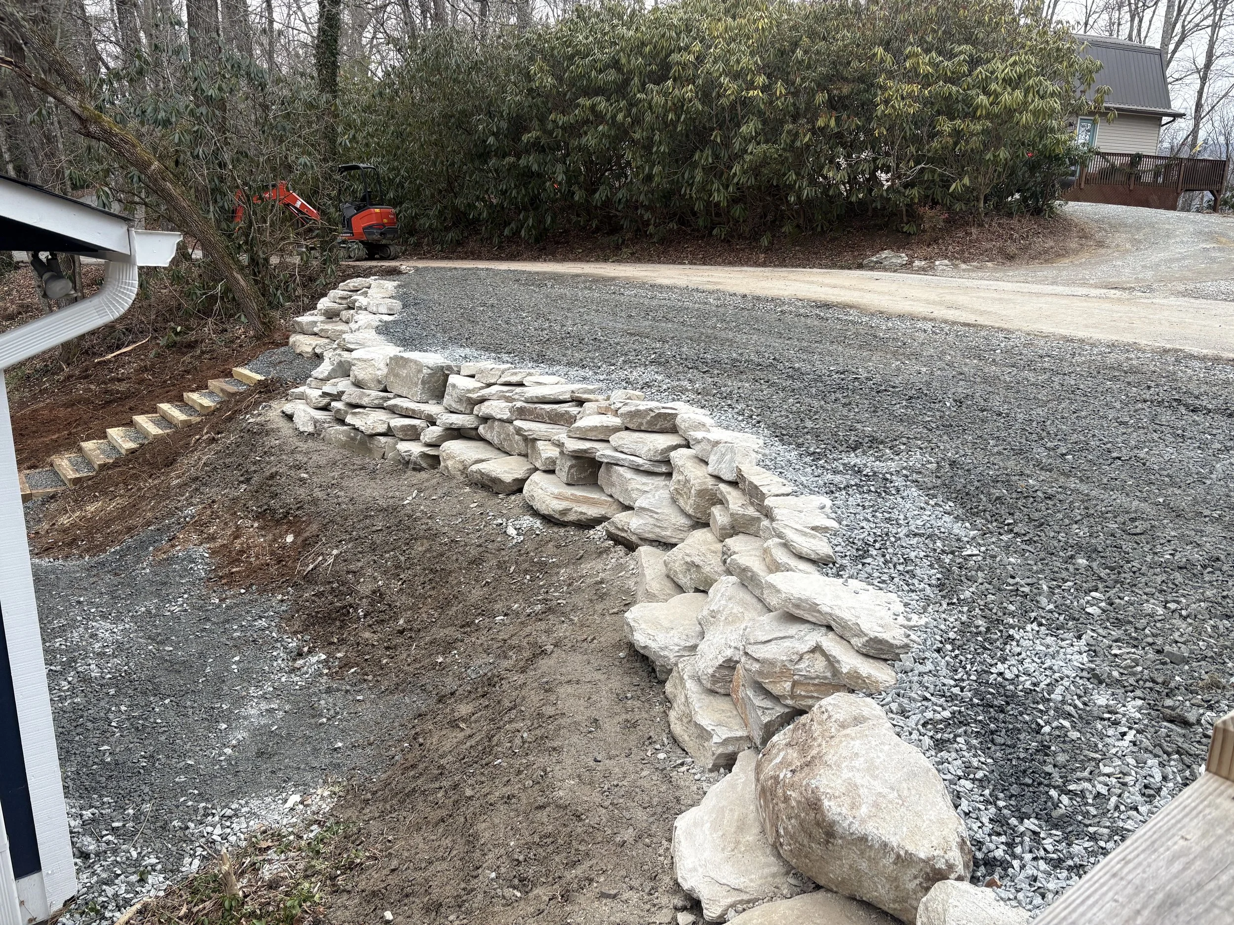 A stone retaining wall under construction along a dirt and gravel driveway in a residential area. A red piece of equipment is visible in the background near bushes and trees.