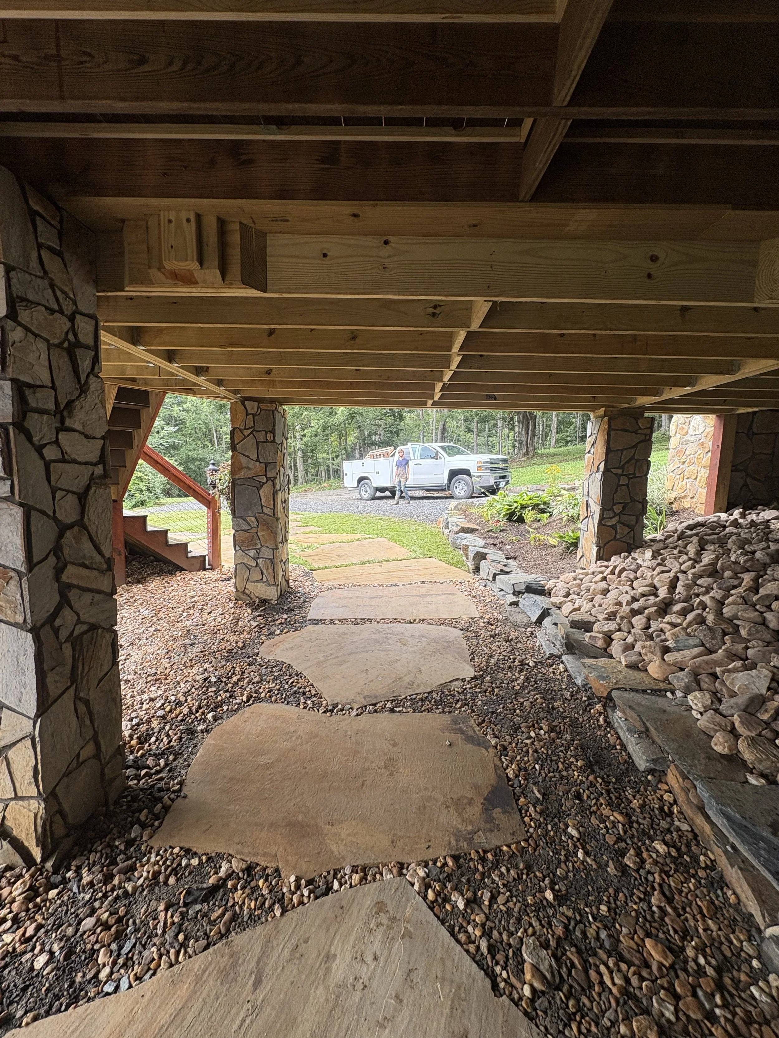 Underneath a wooden deck with stone columns, a stone pathway leads to a yard with a truck and a person standing next to it, surrounded by greenery and trees.