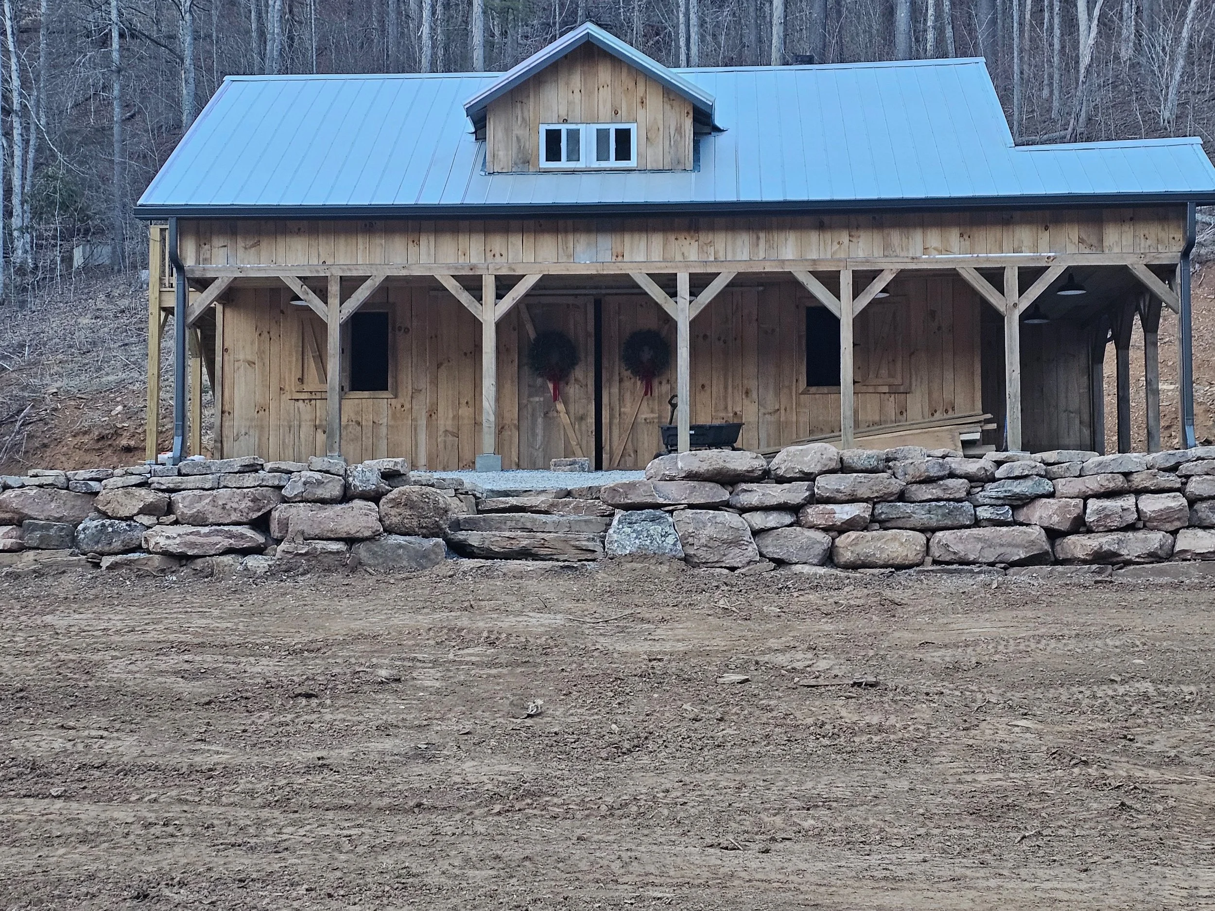 A wooden house under construction with a metal roof, surrounded by trees and a stone retaining wall.