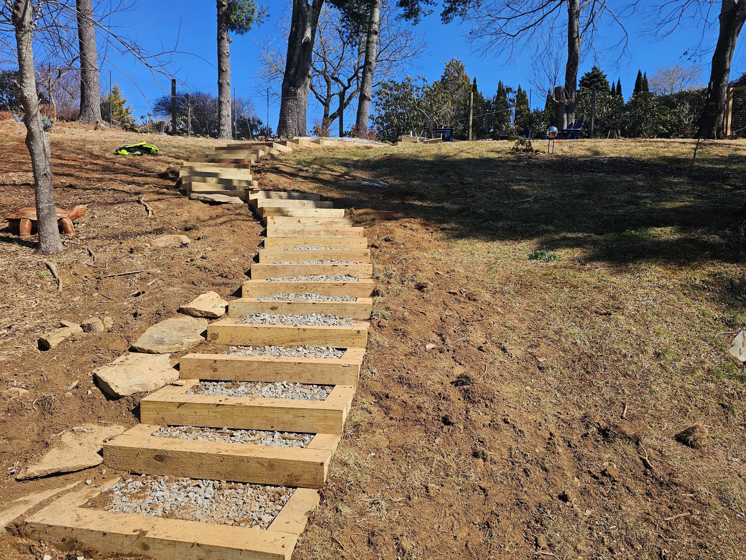 Wooden steps forming a staircase on a sloped outdoor dirt hillside with trees and a blue sky in the background.