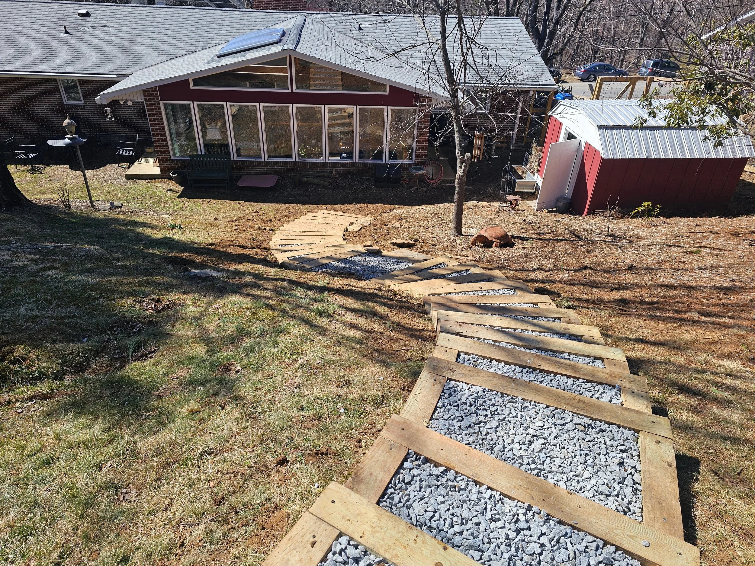 Backyard with a newly constructed gravel and wooden path winding down a grassy and patchy dirt slope towards a brick house. There is a red shed with a metal roof, leafless trees, and parked cars in the background.