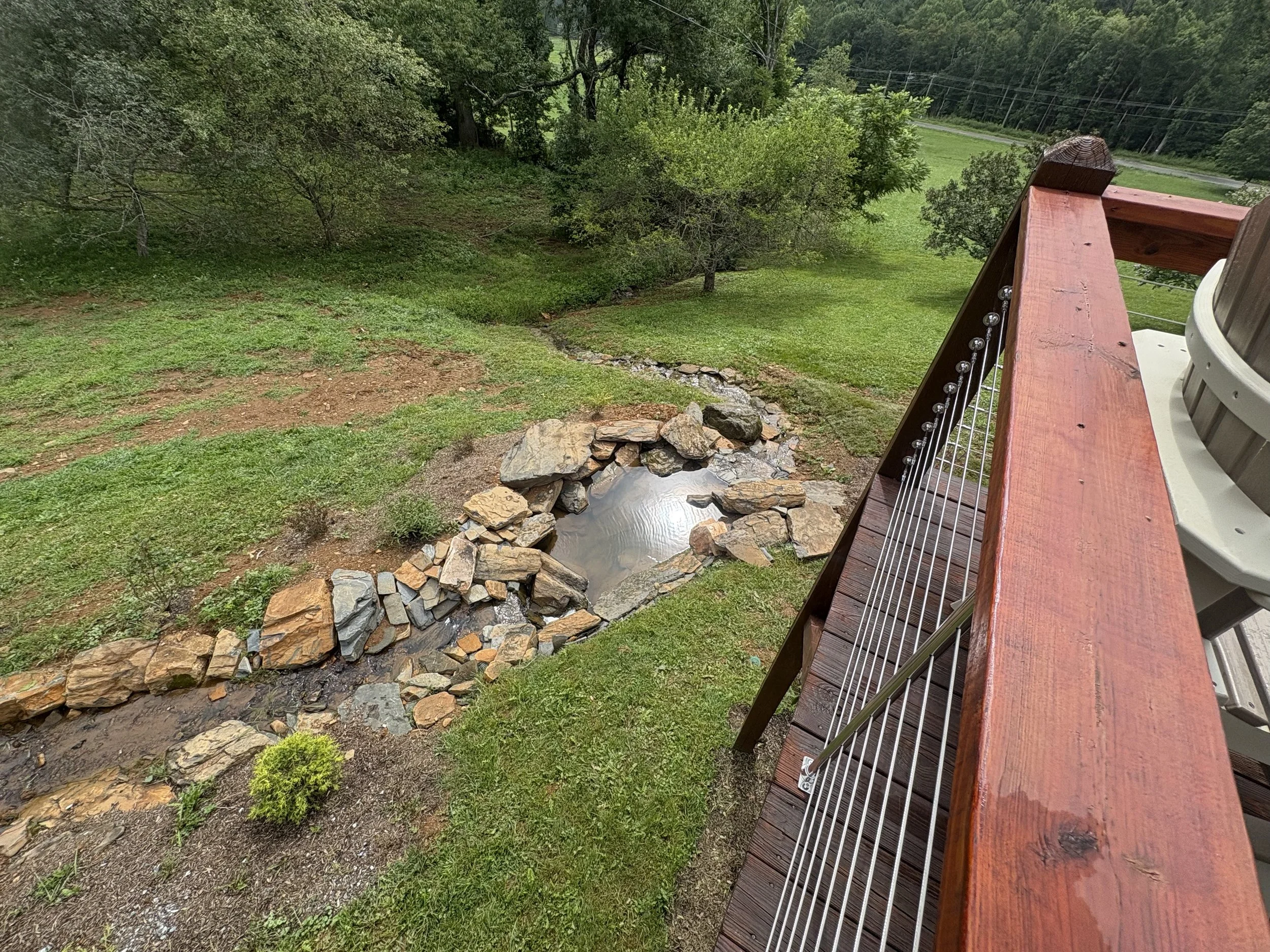 View from a deck showing a small stream with rocks, surrounding green grass, trees, and power lines in the distance.