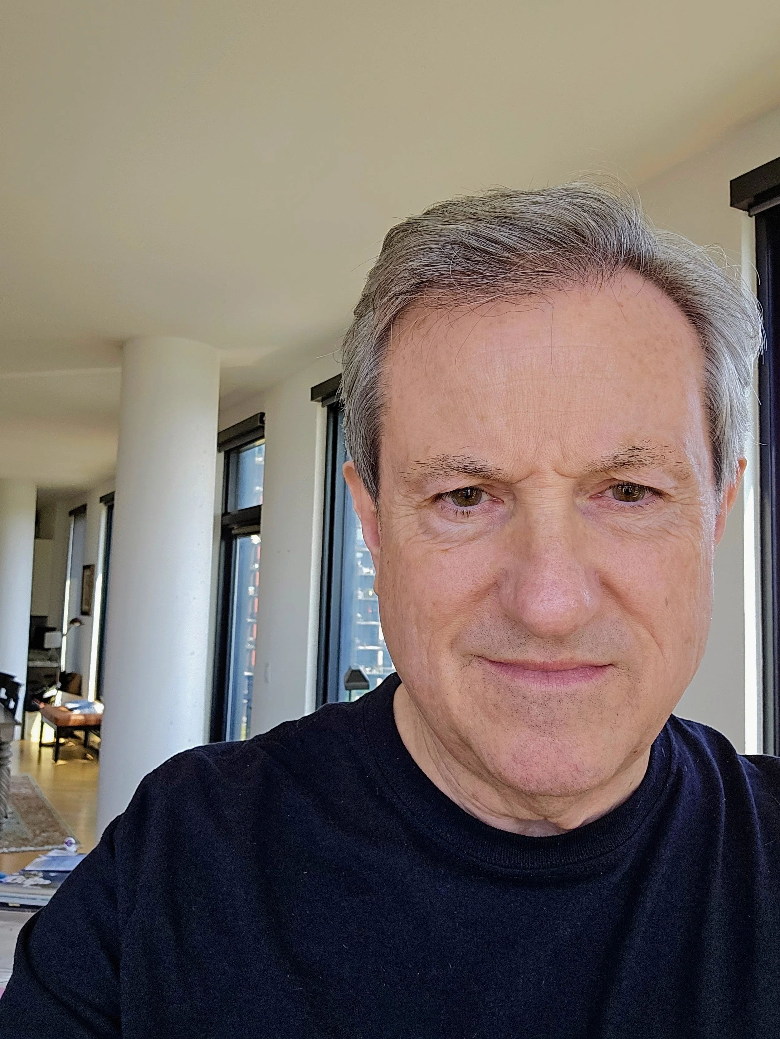 Close-up of a middle-aged man with gray hair wearing a black shirt, indoors with large windows and a city view in the background.