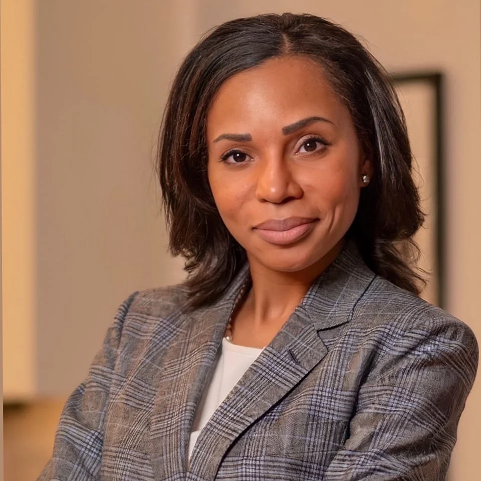 A professional woman with shoulder-length dark hair wearing a gray plaid blazer smiling at the camera.