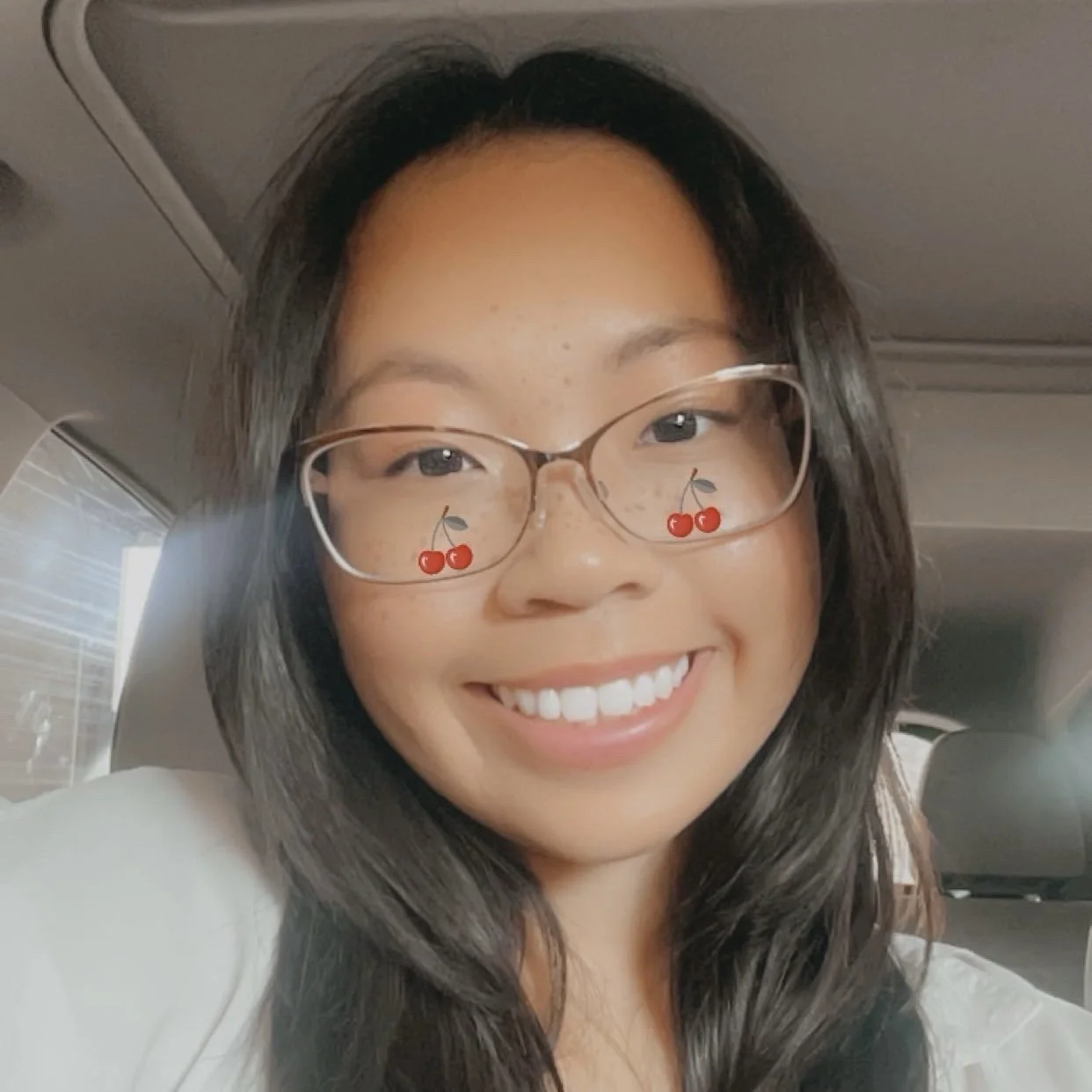 A young woman with glasses smiling inside a vehicle, with a cherry emoji filter on her glasses.