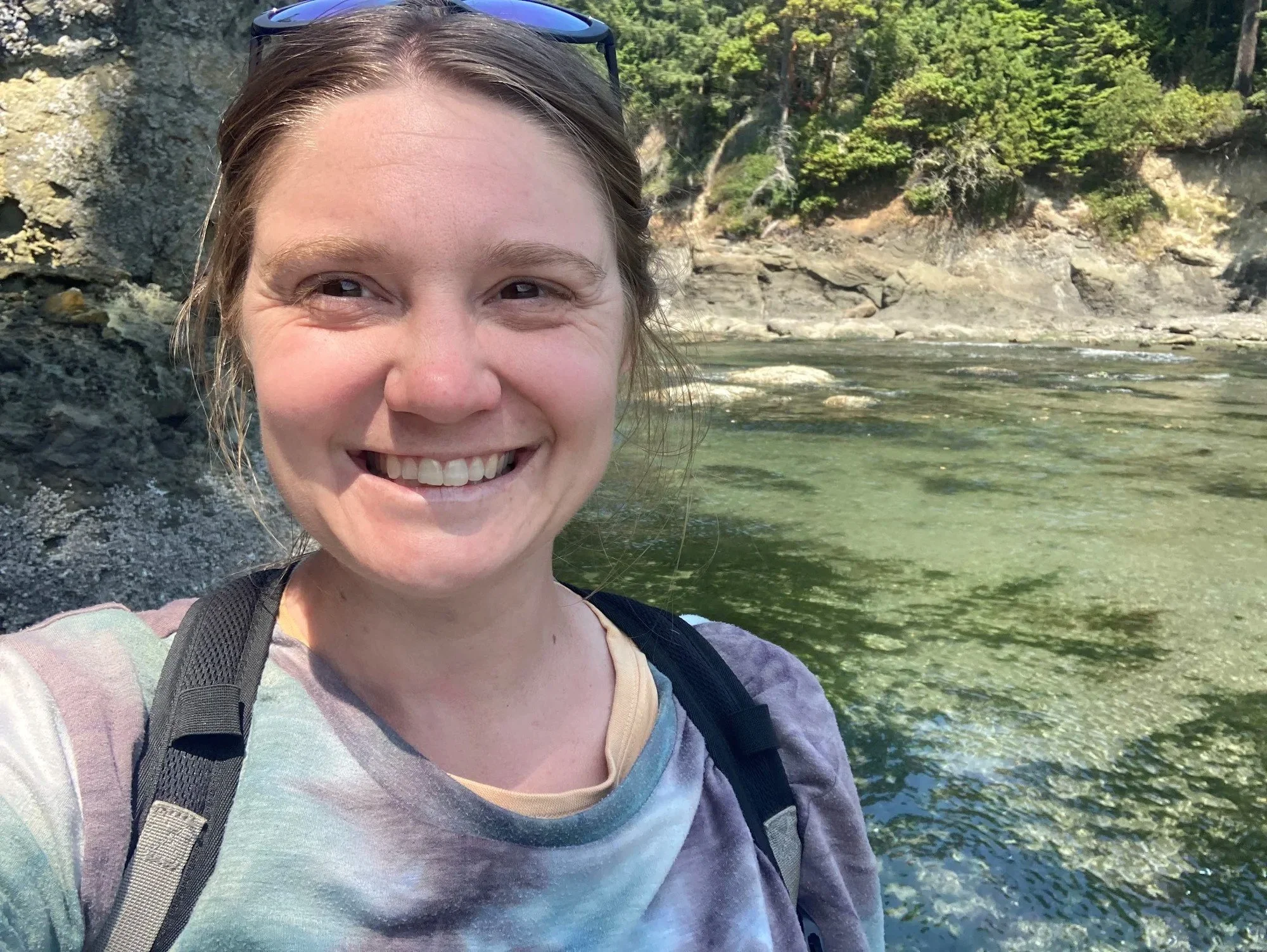 A smiling woman with sunglasses on her head, standing near a river in a wooded area.