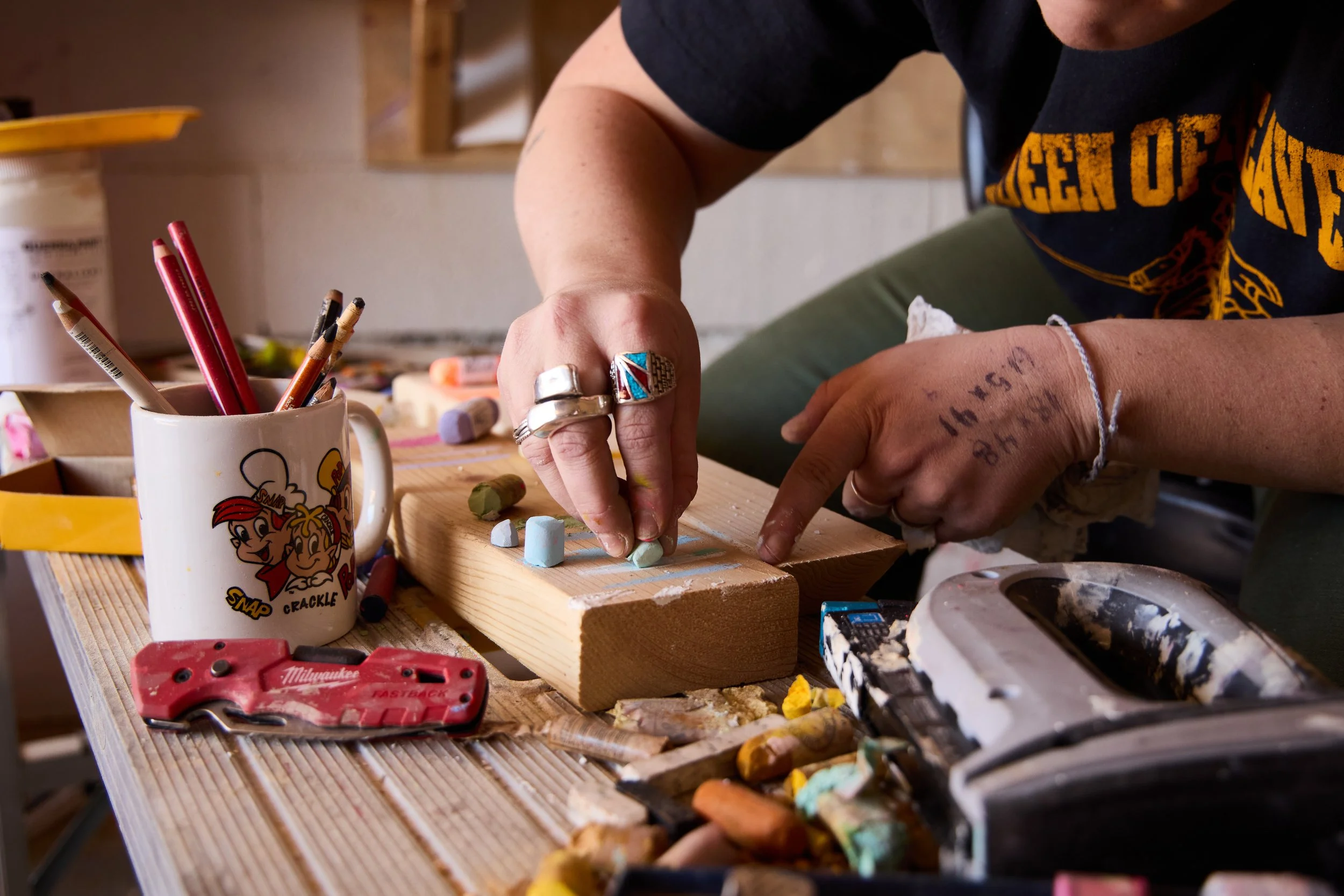Person's hands carving chalk sculptures on a wooden block surrounded by art supplies and chalk pieces, with a coffee mug featuring cartoon characters and a utility knife nearby.